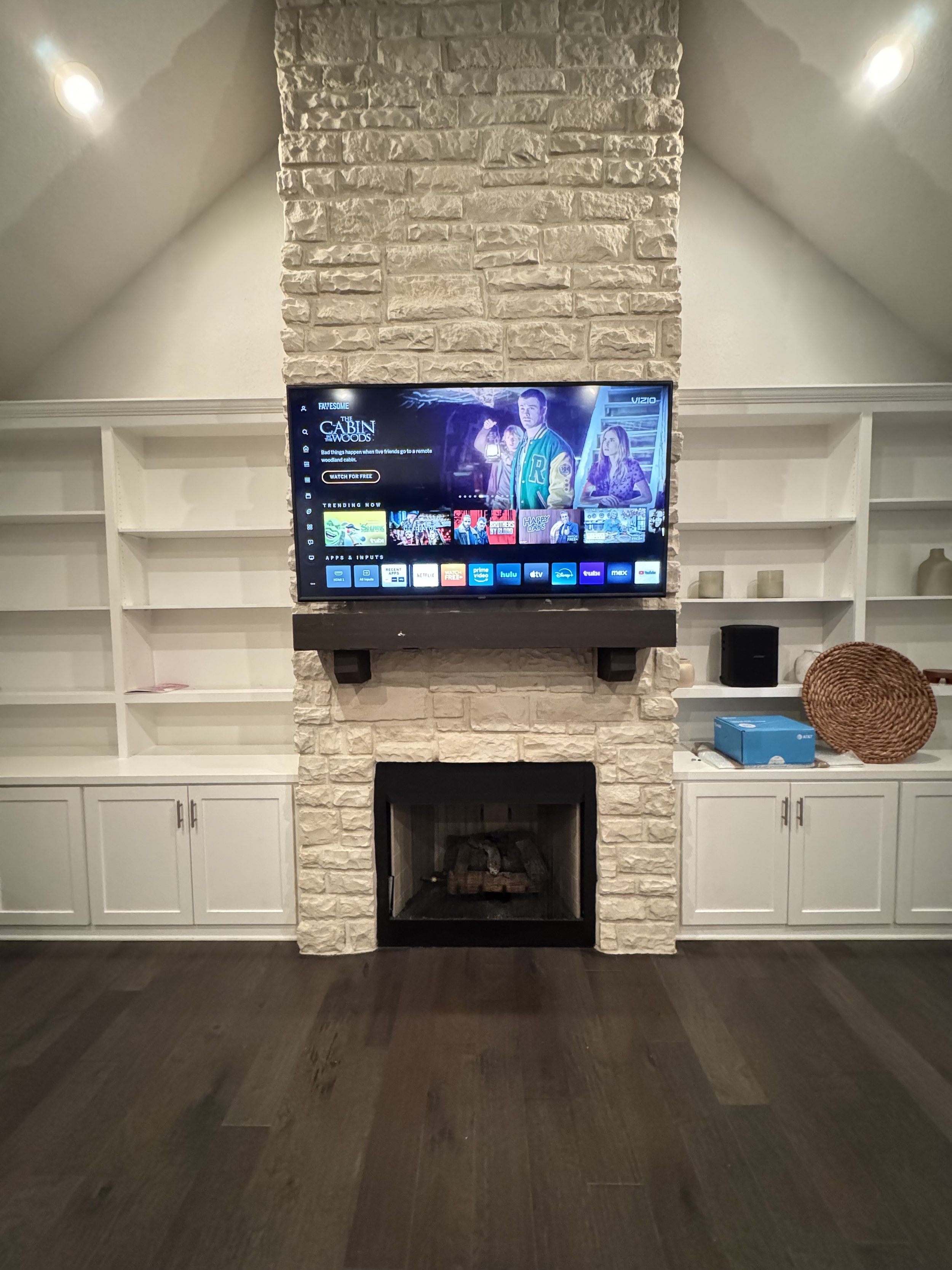 Living room with a stone fireplace, mounted TV, and built-in white shelving on an angled ceiling. The TV shows a streaming app interface, and the fireplace has logs inside. There are small decorative items on the shelves, a blue box, and a woven bask