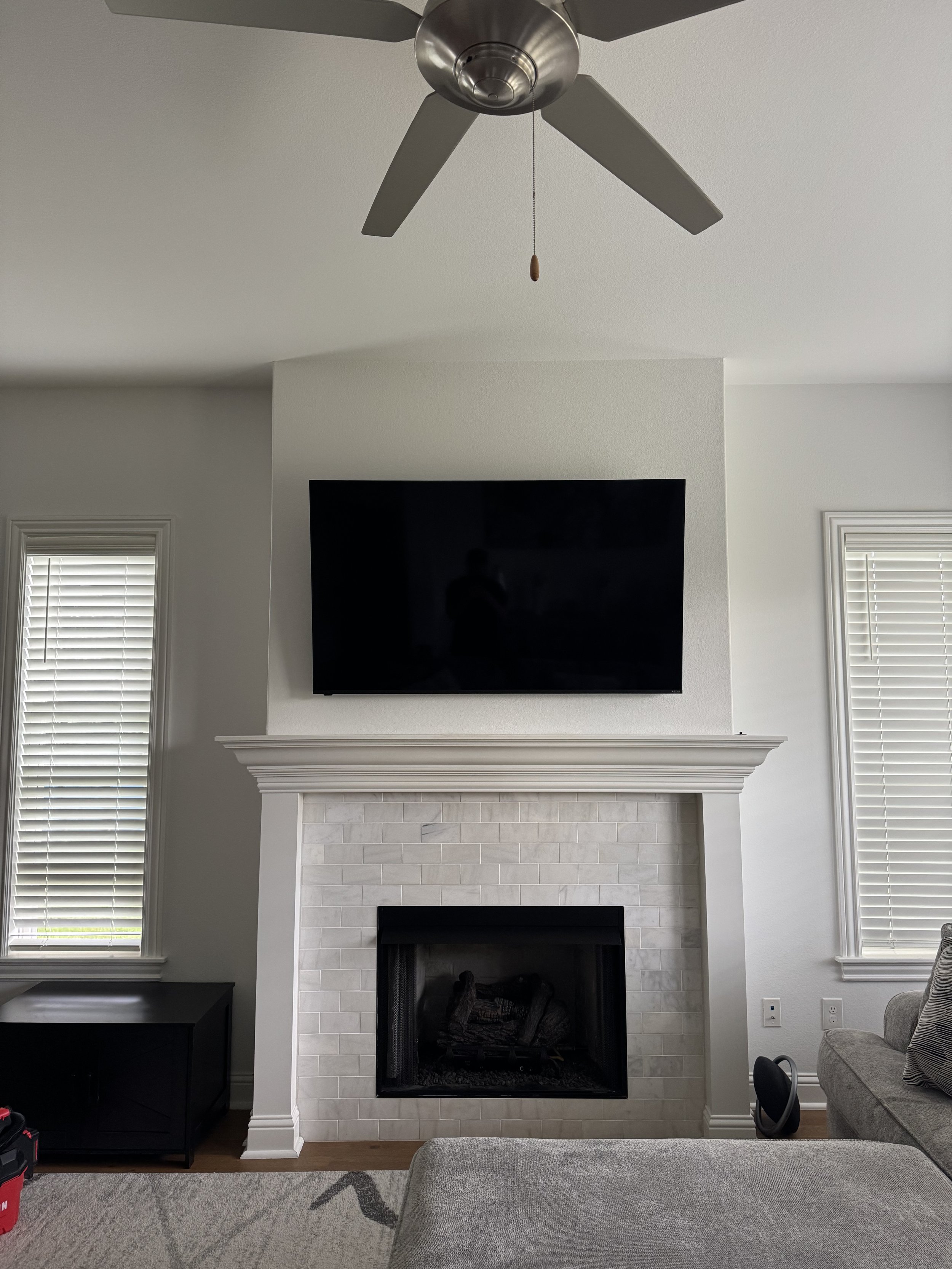 Living room fireplace with a mounted flat-screen TV above it, two windows with blinds on either side, and a ceiling fan overhead.