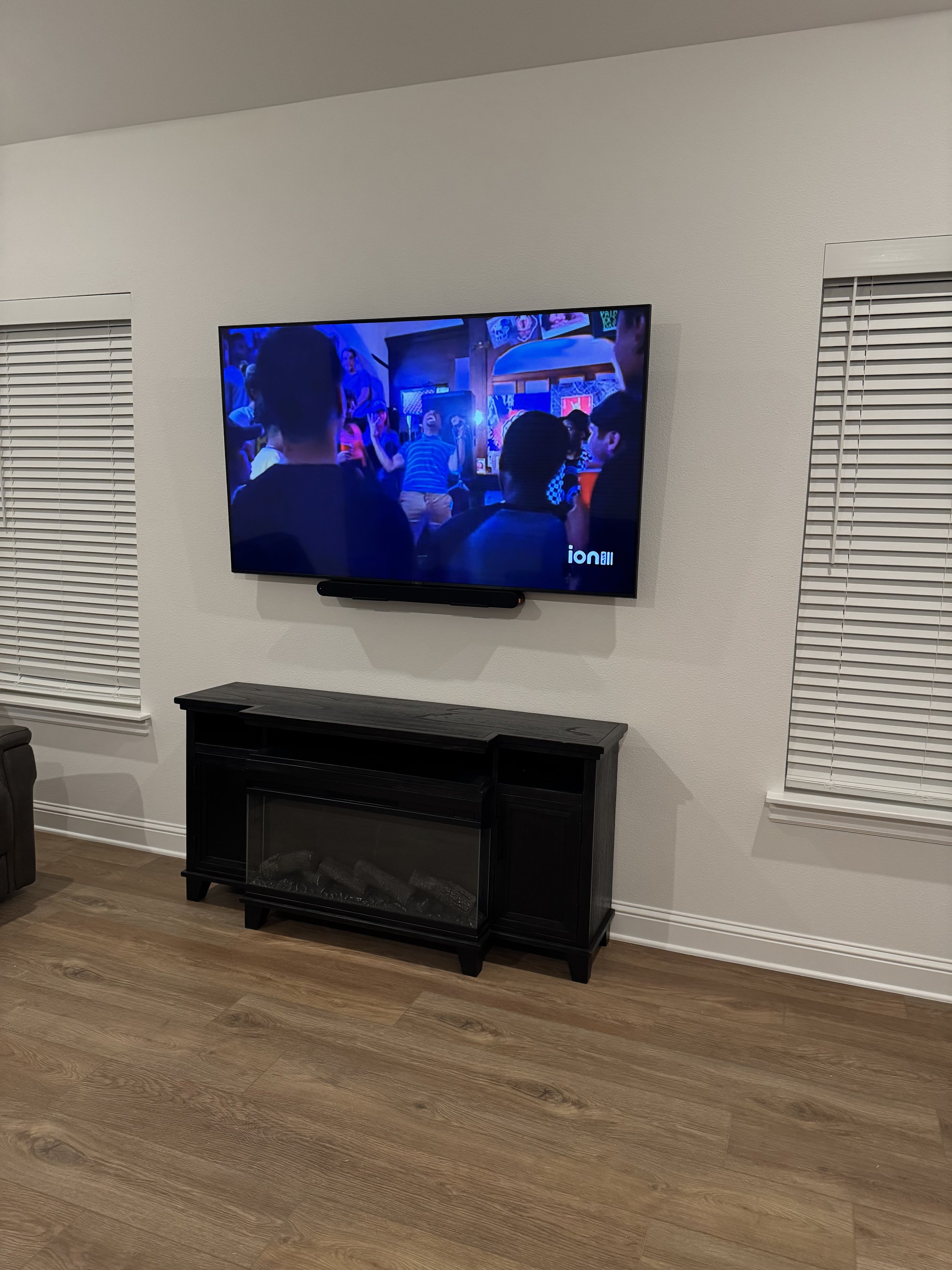 Living room with wall-mounted TV displaying a crowd scene at an indoor event, black entertainment center below the TV, light-colored hardwood floor, white walls, two windows with closed blinds.