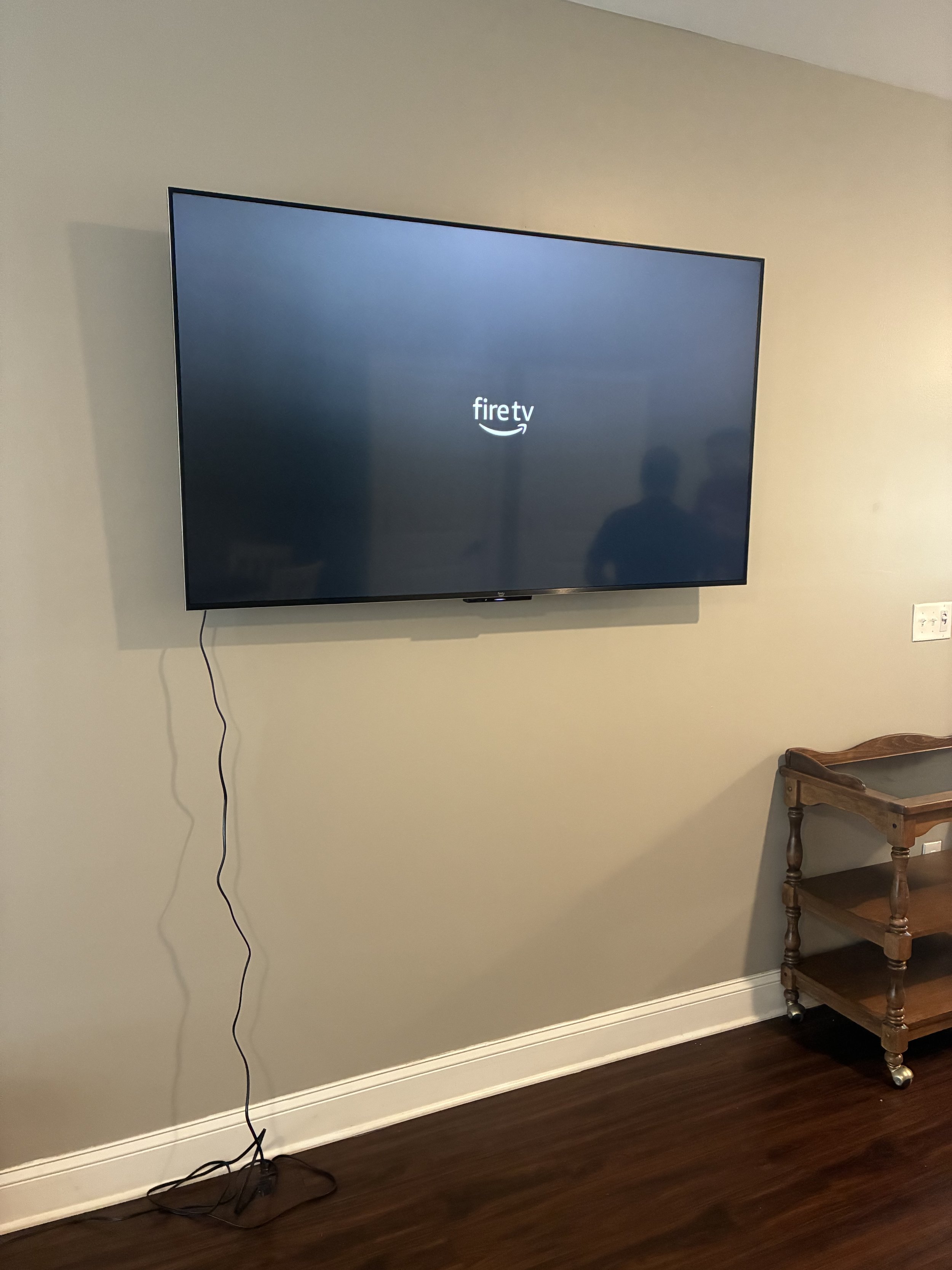A flat-screen TV mounted on a beige wall displaying the 'fire tv' logo, with a black power cord hanging down to the floor and a small wooden table nearby.