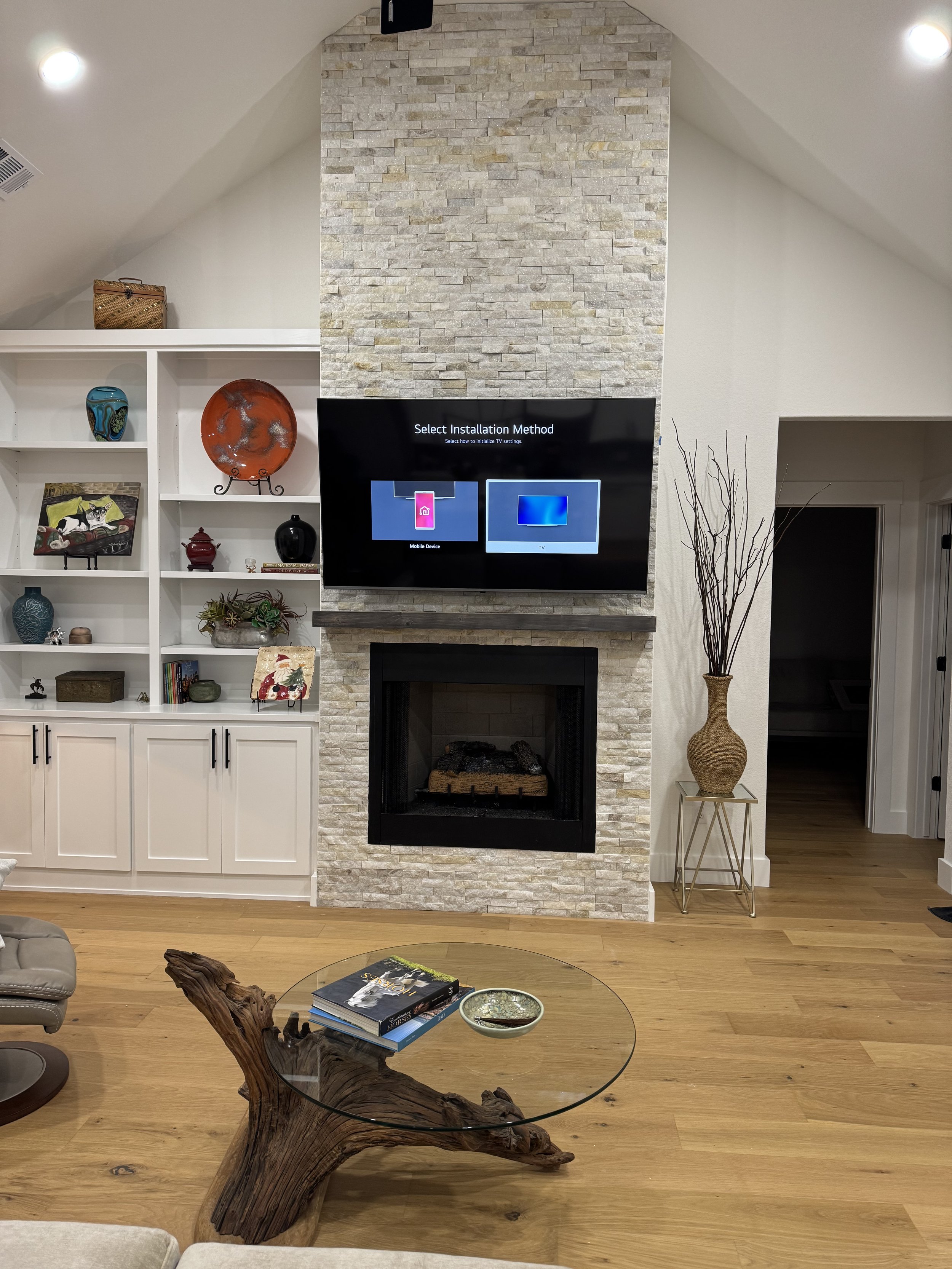 Living room with a stone fireplace, wall-mounted TV displaying installation method, white built-in shelves with decorative items, a tall vase with branches, and a glass coffee table with magazines and a bowl.