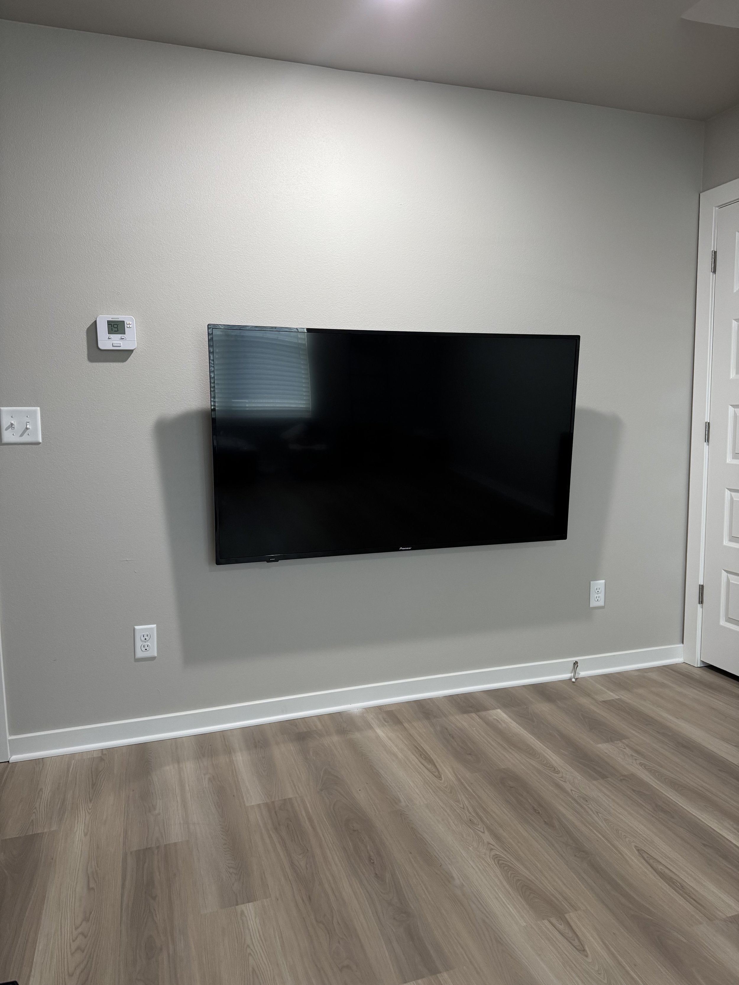 Empty living room wall with a mounted flat-screen TV, thermostat, outlets, and light-colored hardwood flooring.