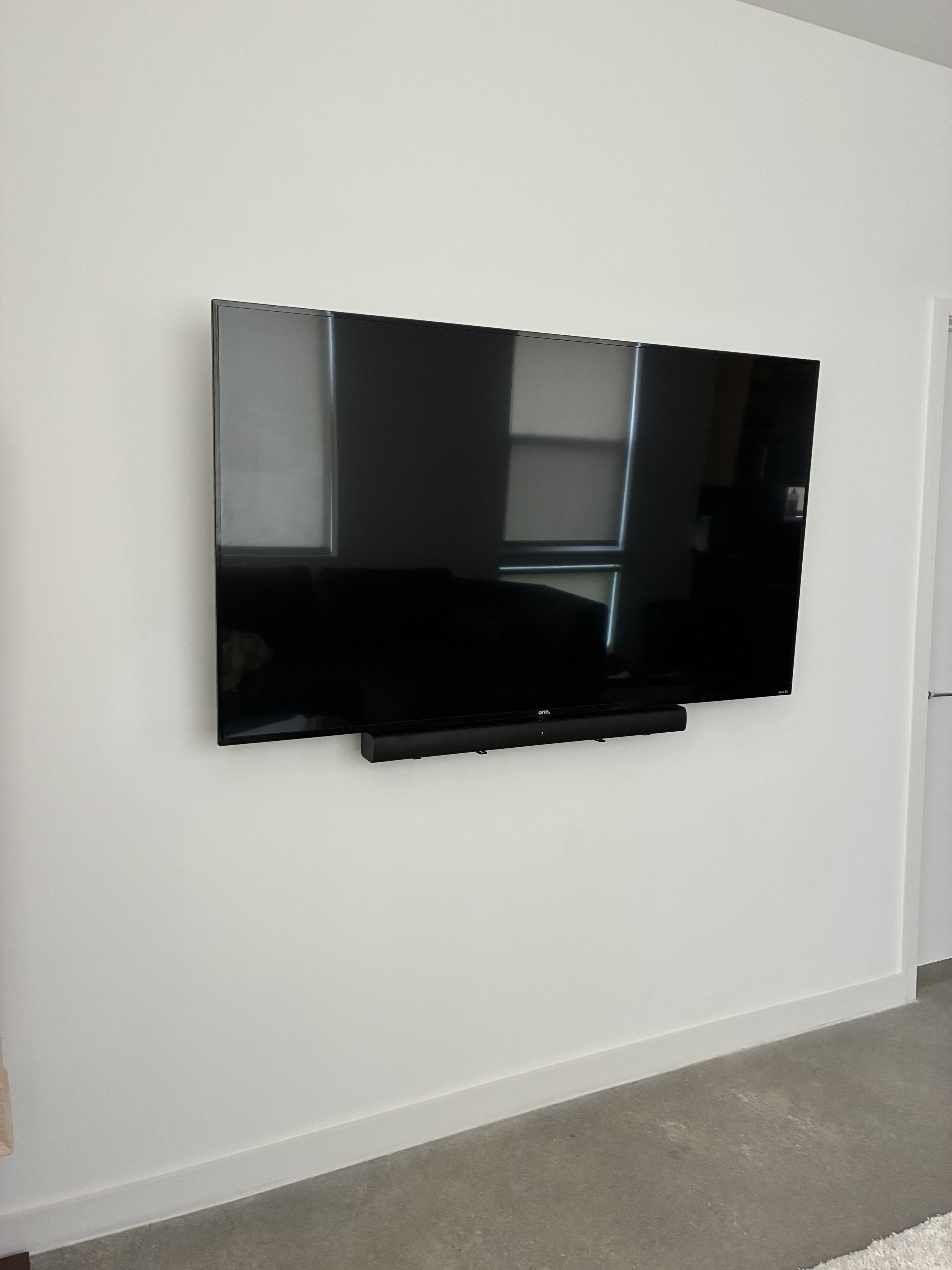 A flat screen TV mounted on a white wall with a soundbar below it, reflecting a window and some furniture.