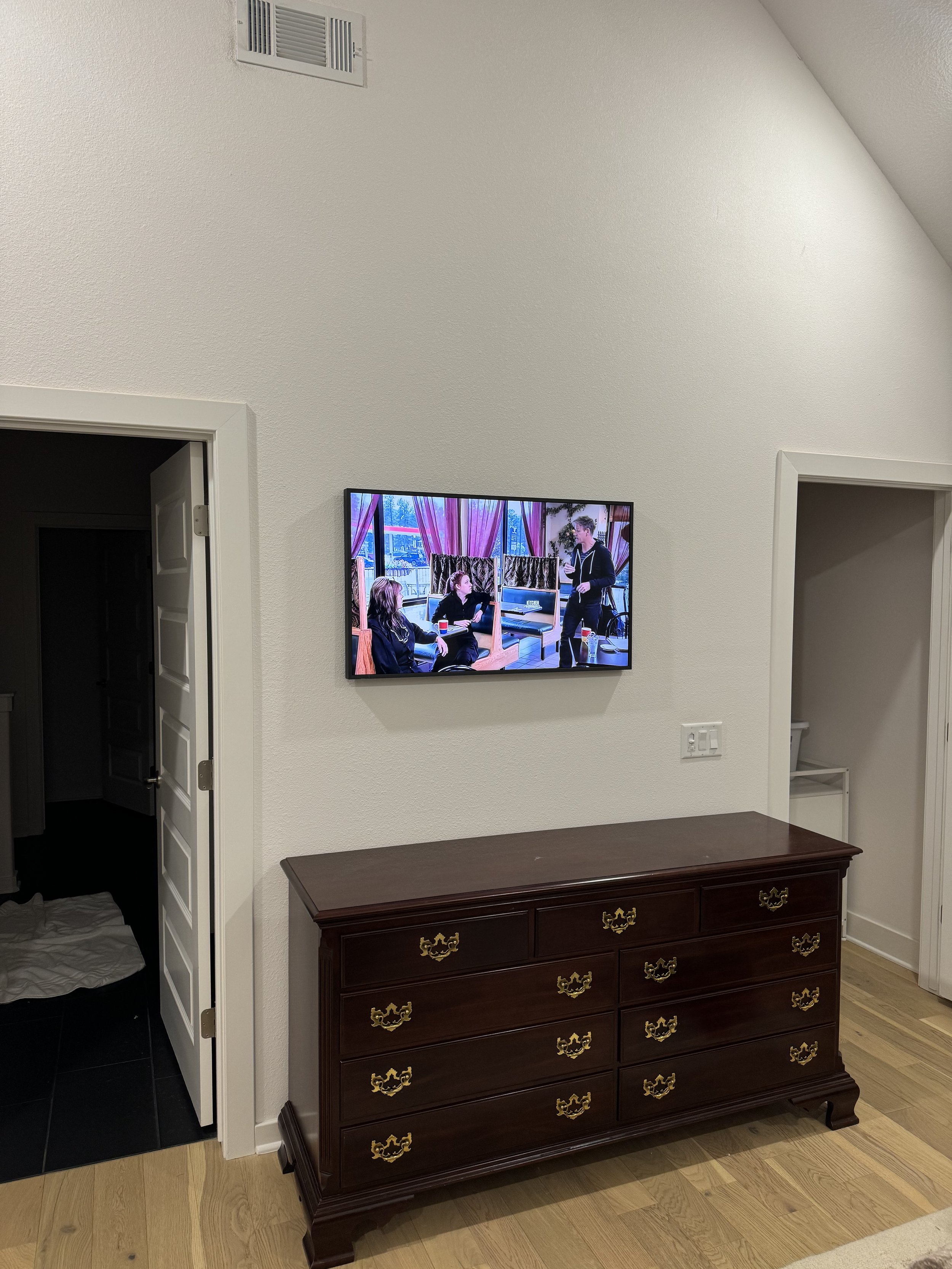 Interior of a room with a wall-mounted TV displaying a scene with people, a dark wooden dresser with gold handles underneath, and two doorways on either side of the dresser.