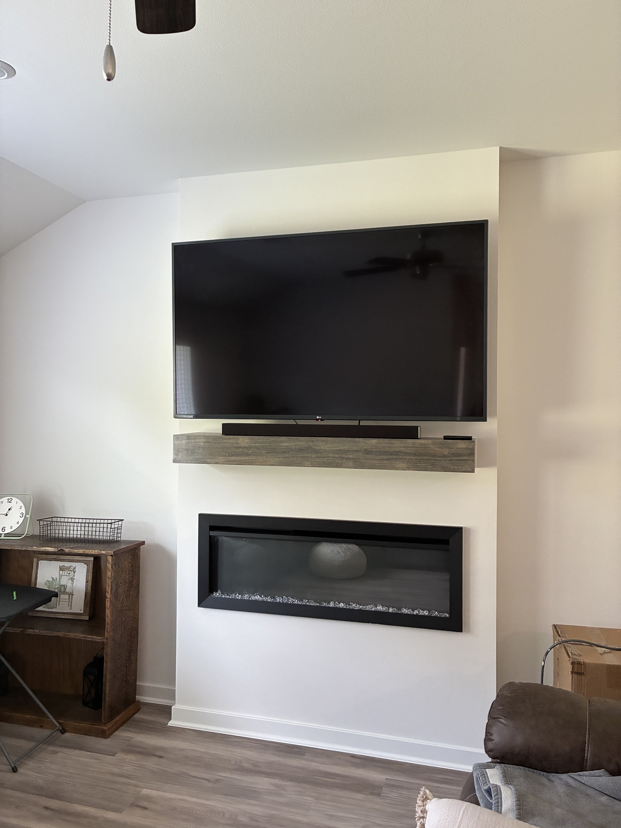 Living room with wall-mounted TV above a fireplace, decorative fireplace with a black frame, and a small wooden cabinet with a clock and framed picture. Part of a brown sofa and a cardboard box are visible.
