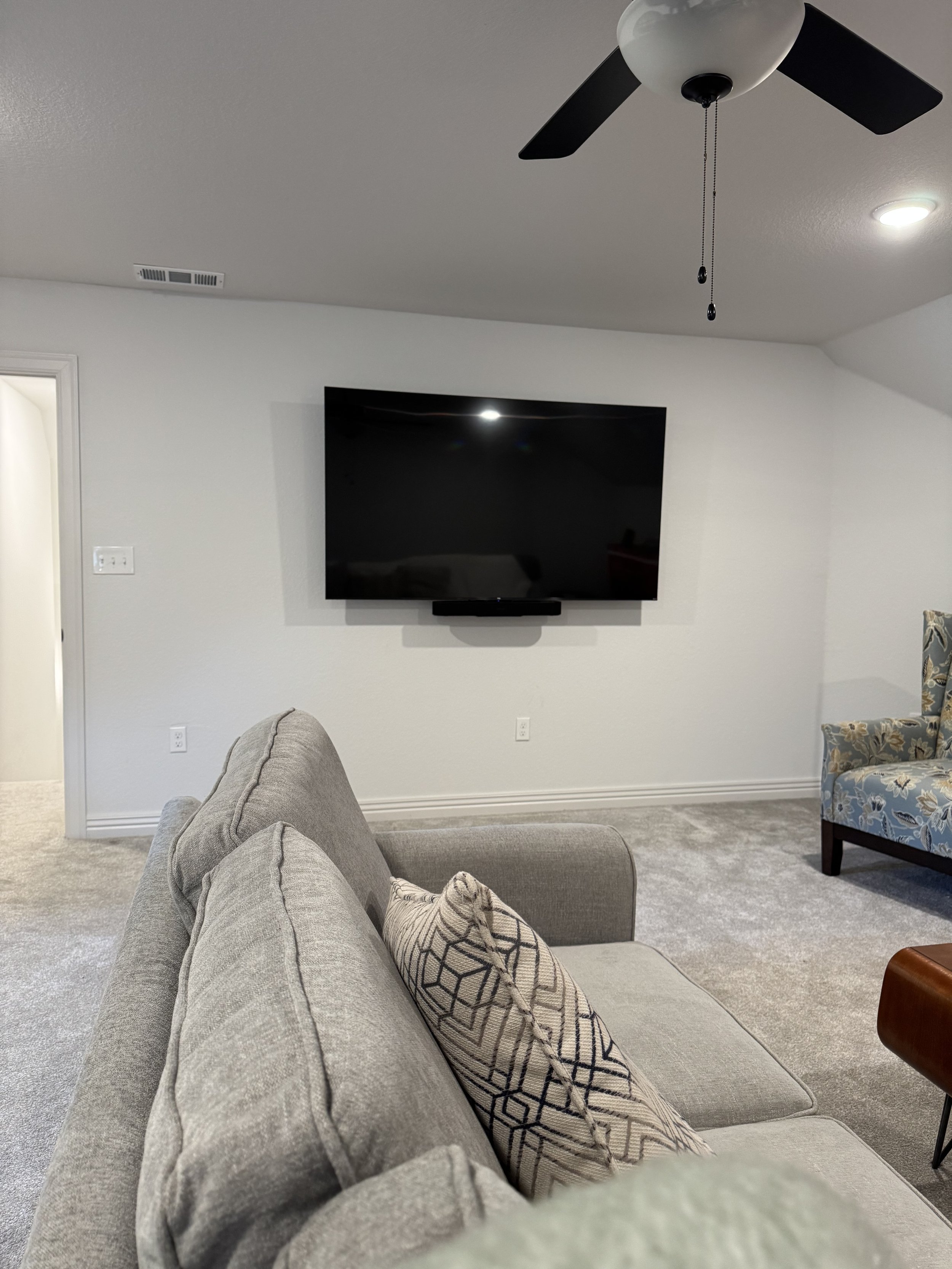 Living room with a mounted flat-screen TV, ceiling fan, gray sofa with patterned pillow, beige carpet, and armchair with floral upholstery.