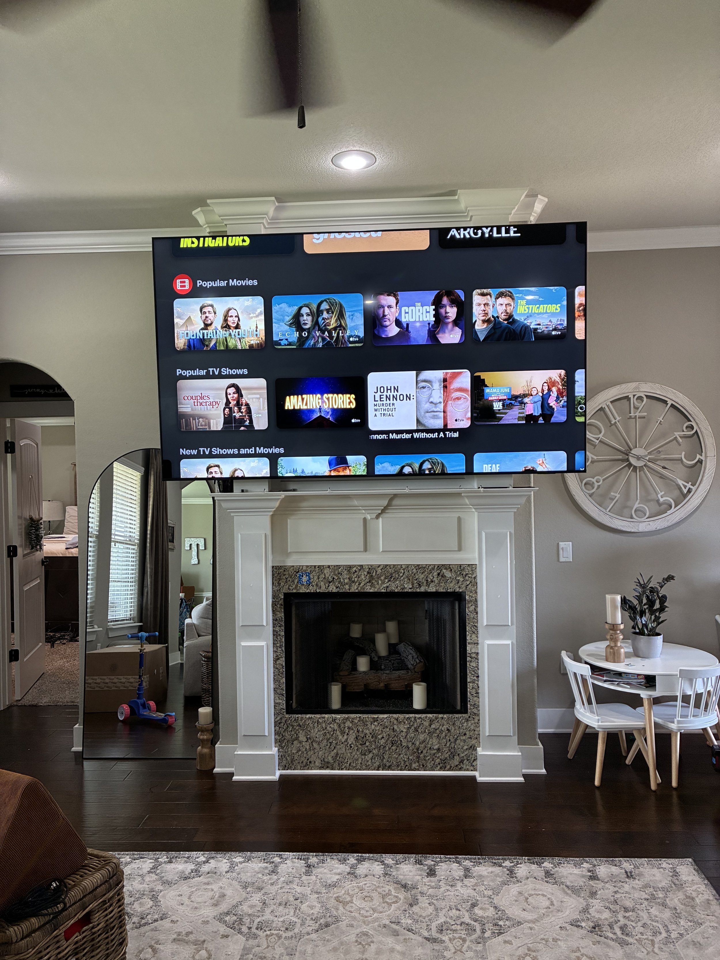 Living room with a large wall-mounted TV displaying a streaming app, white fireplace mantel, decorative candles inside the fireplace, a large round clock on the wall, a small white table with chairs, a basket, a bunny-shaped scooter, and a mirror ref