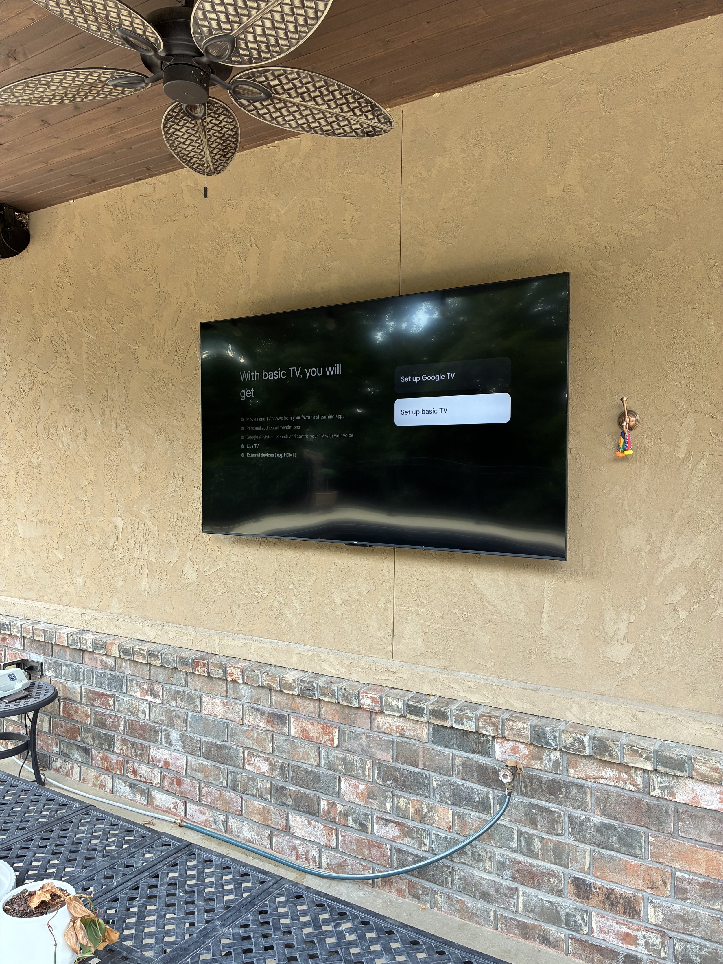Outdoor wall with a mounted flat-screen TV displaying setup options for a basic TV or Google TV, and a ceiling fan with leaf-shaped blades.