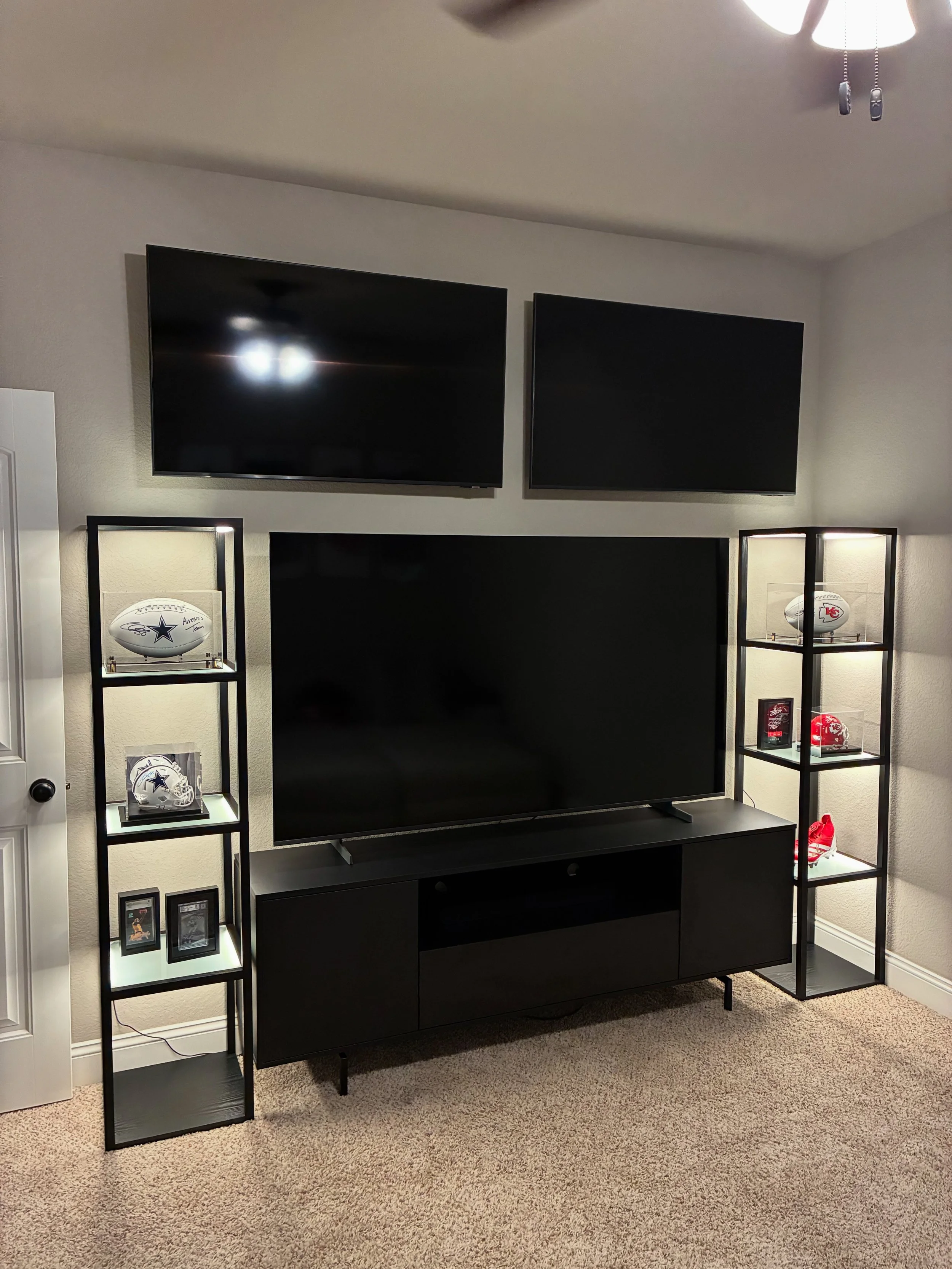 Living room entertainment center with a black TV on a black stand, two display shelves on each side with football memorabilia and photos, and a ceiling fan.