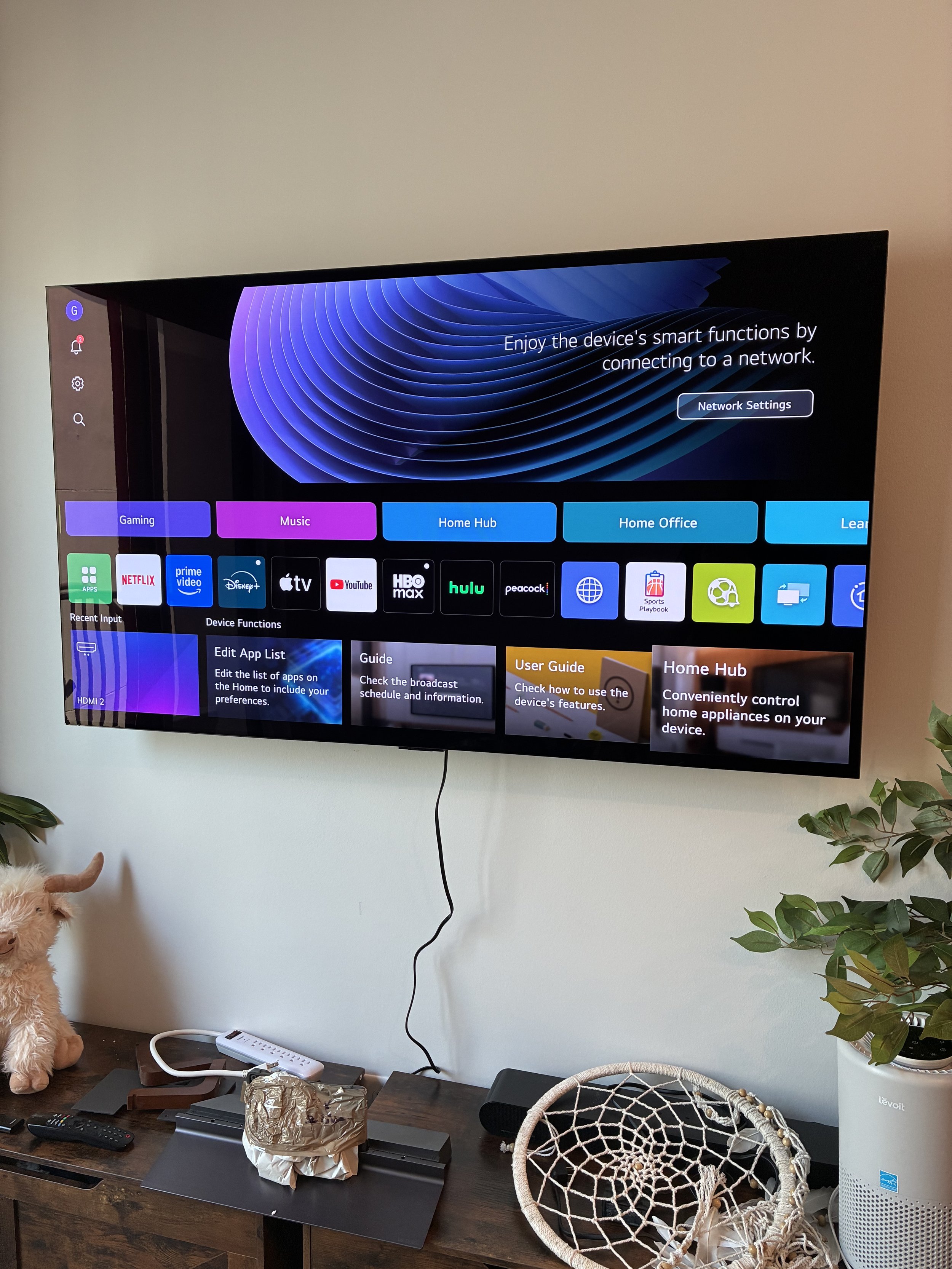 Wall-mounted flat-screen TV displaying a streaming device menu with options such as Gaming, Music, Home Hub, and Home Office. Below the TV, there is a wooden table with various items including a stuffed llama, an extension cord, a wrapped foil object