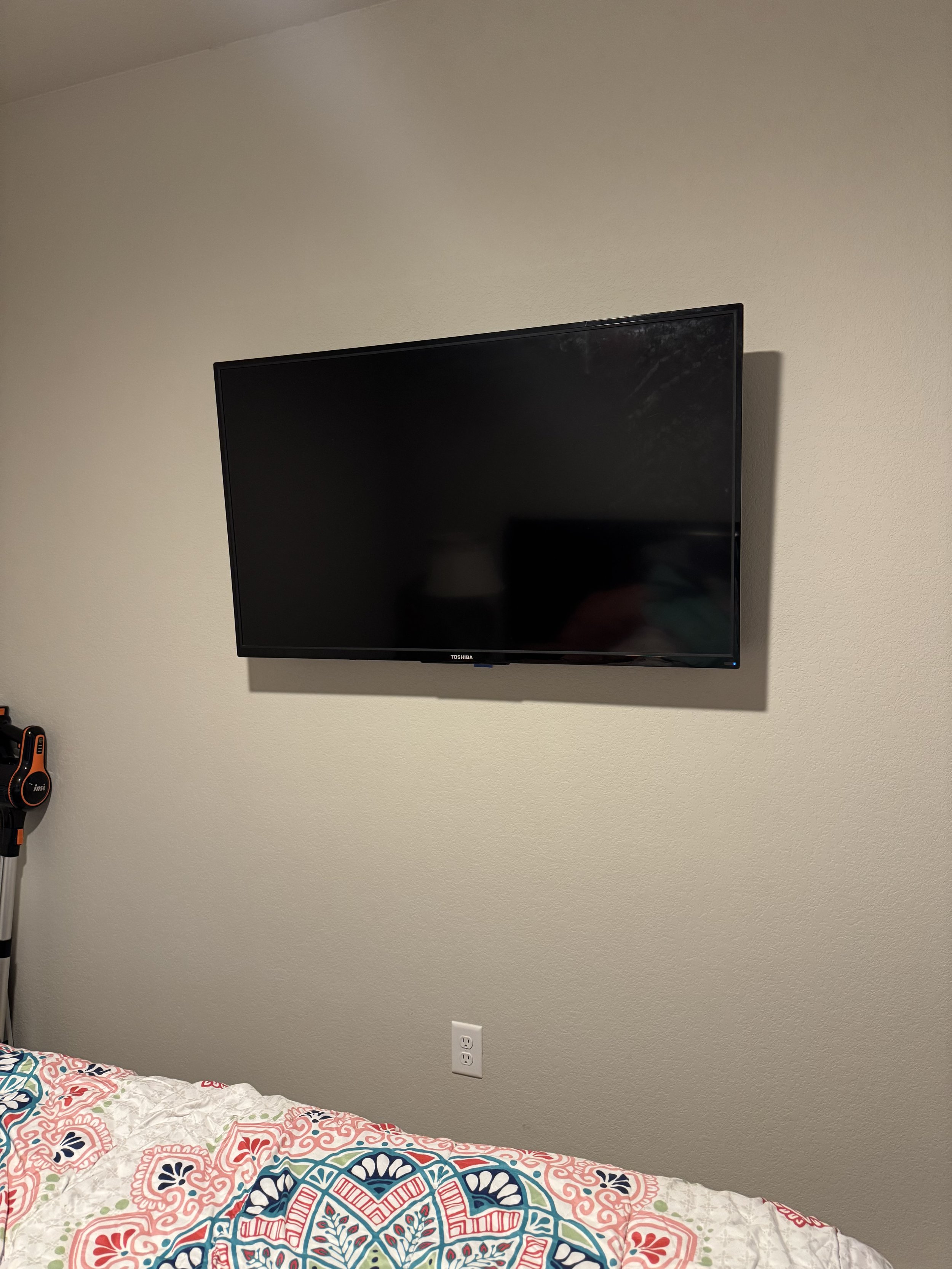 A flat-screen Toshiba television mounted on a plain beige wall in a bedroom, with a colorful patterned bedspread in the foreground.
