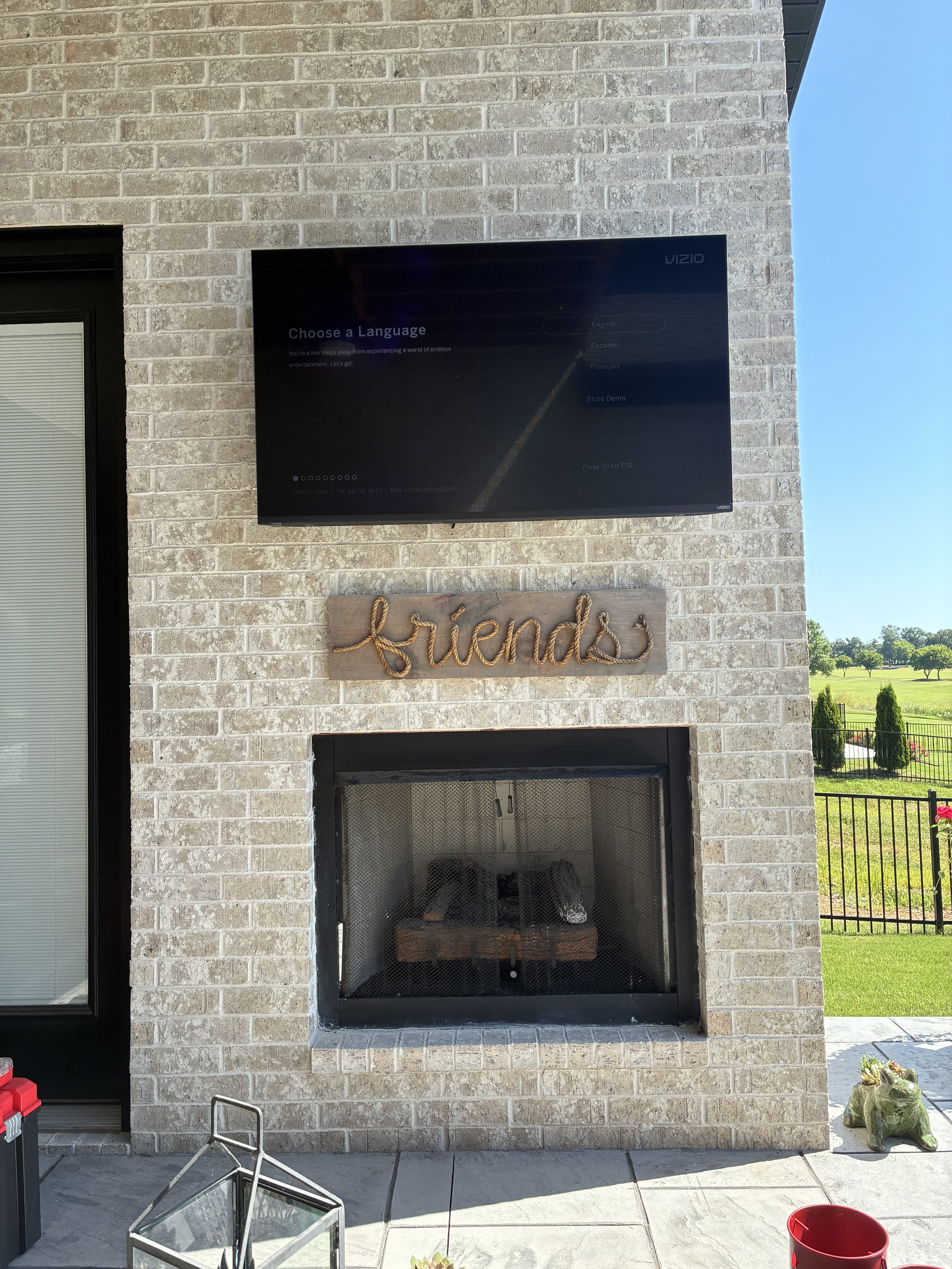 Outdoor brick fireplace with a sign saying 'friends' made of rope, a mounted television displaying a language selection screen, and a green decorative frog sculpture on the patio.