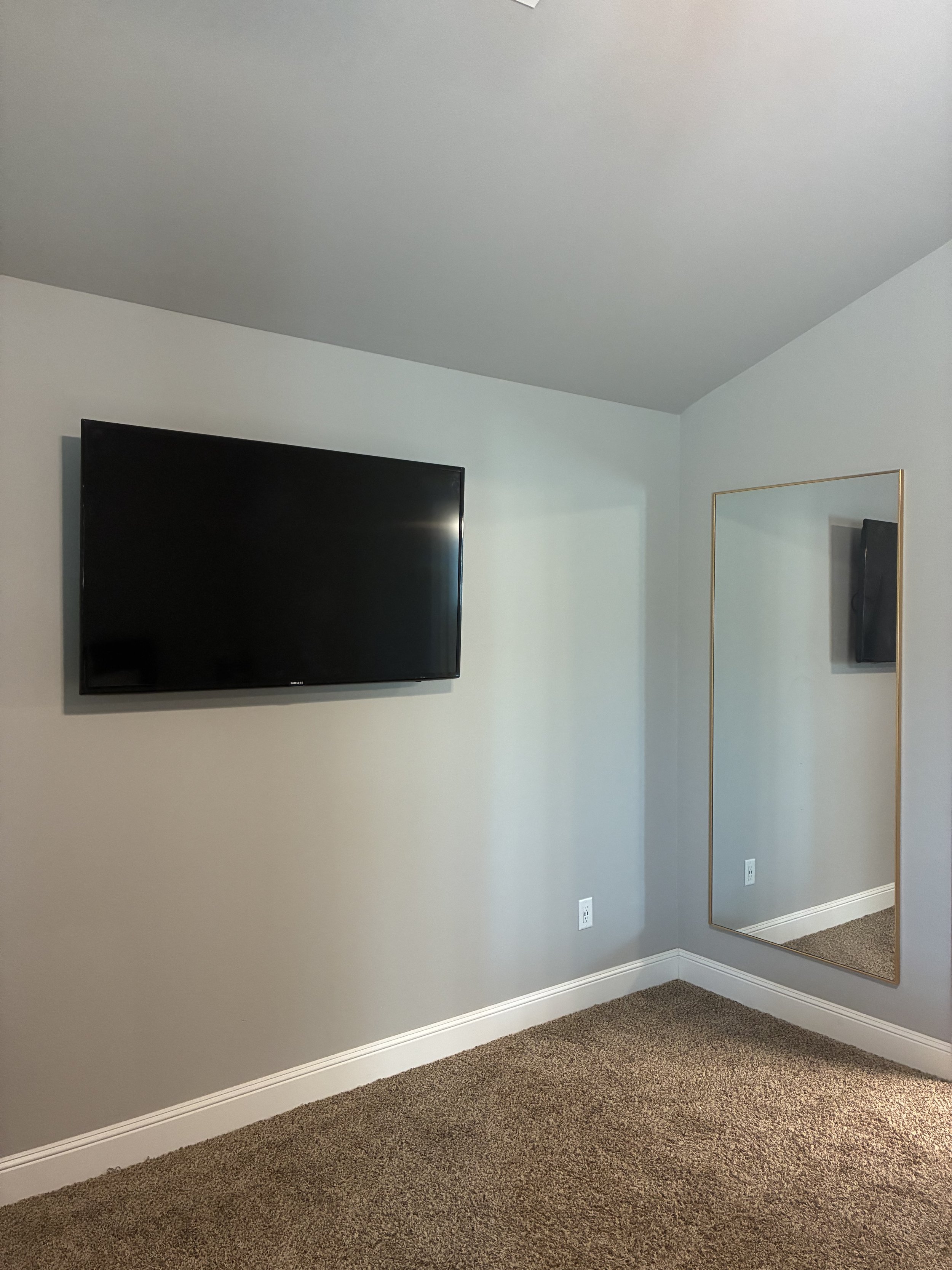 A bedroom corner with a mounted flat-screen TV, a tall rectangular mirror, gray walls, white baseboards, and brown carpet.