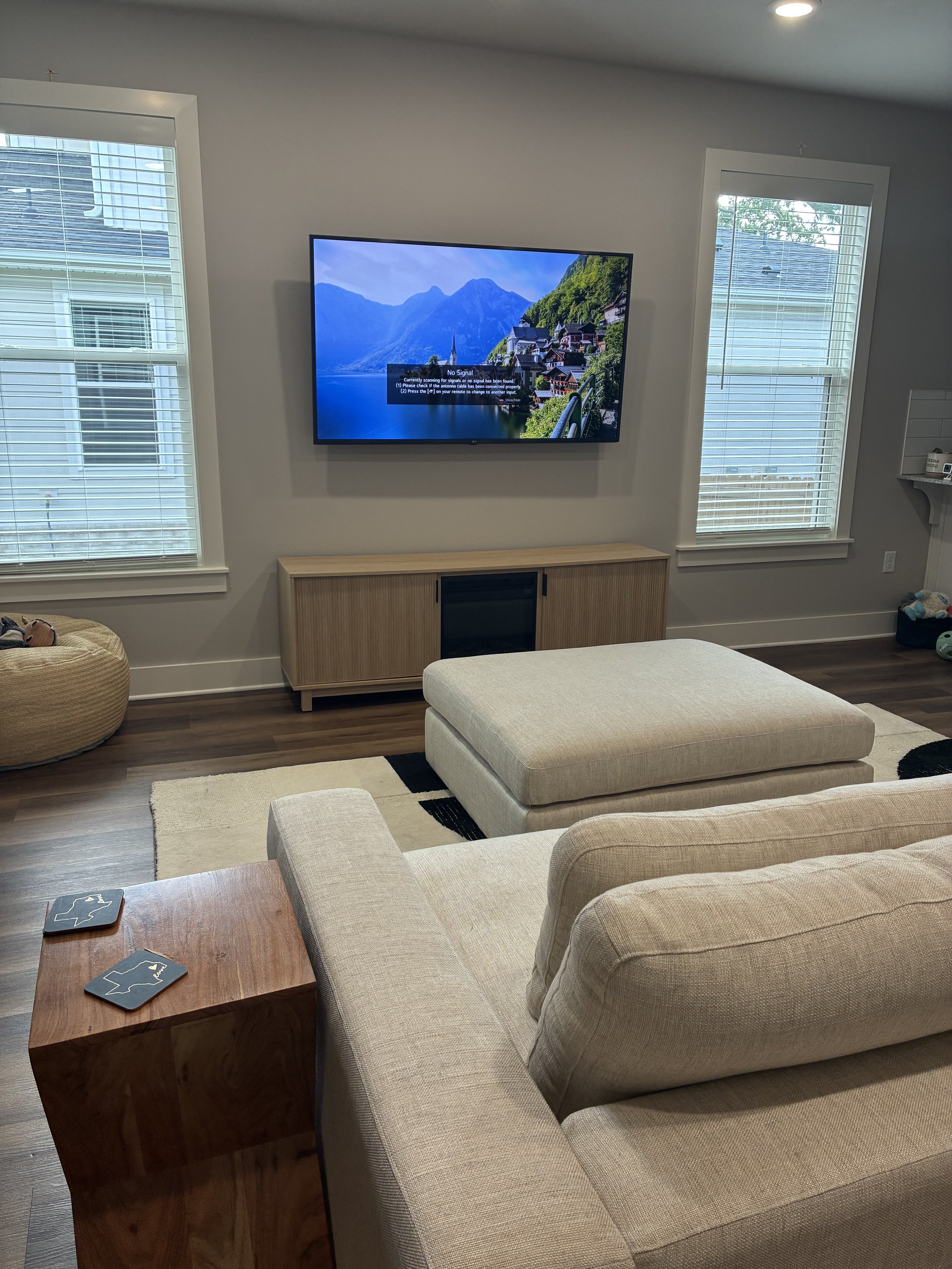 Living room with beige sofa, matching ottoman, wooden coffee table with two coasters, and a wall-mounted TV displaying a mountain lake scene, flanked by two windows with white blinds, and a wooden media console below the TV.