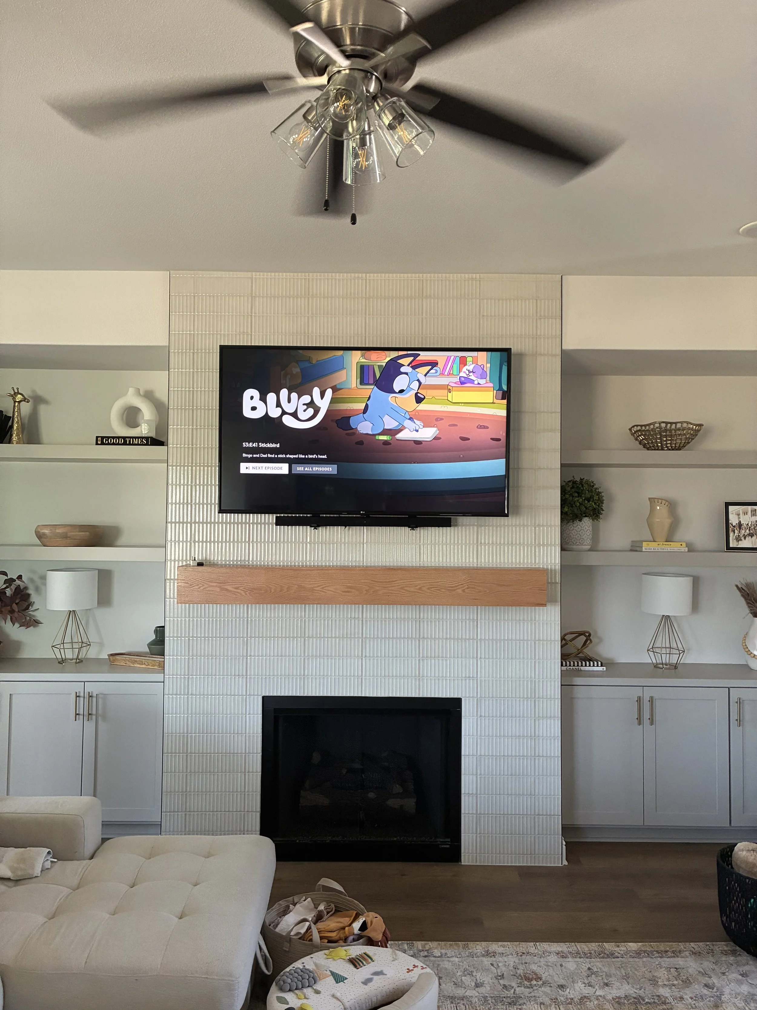 Living room with a wall-mounted TV displaying a blue cartoon dog named Bluey, above a fireplace with a wooden mantel. There are built-in shelves on either side of the fireplace, decorated with plants, vases, pictures, and decorative objects. A white 