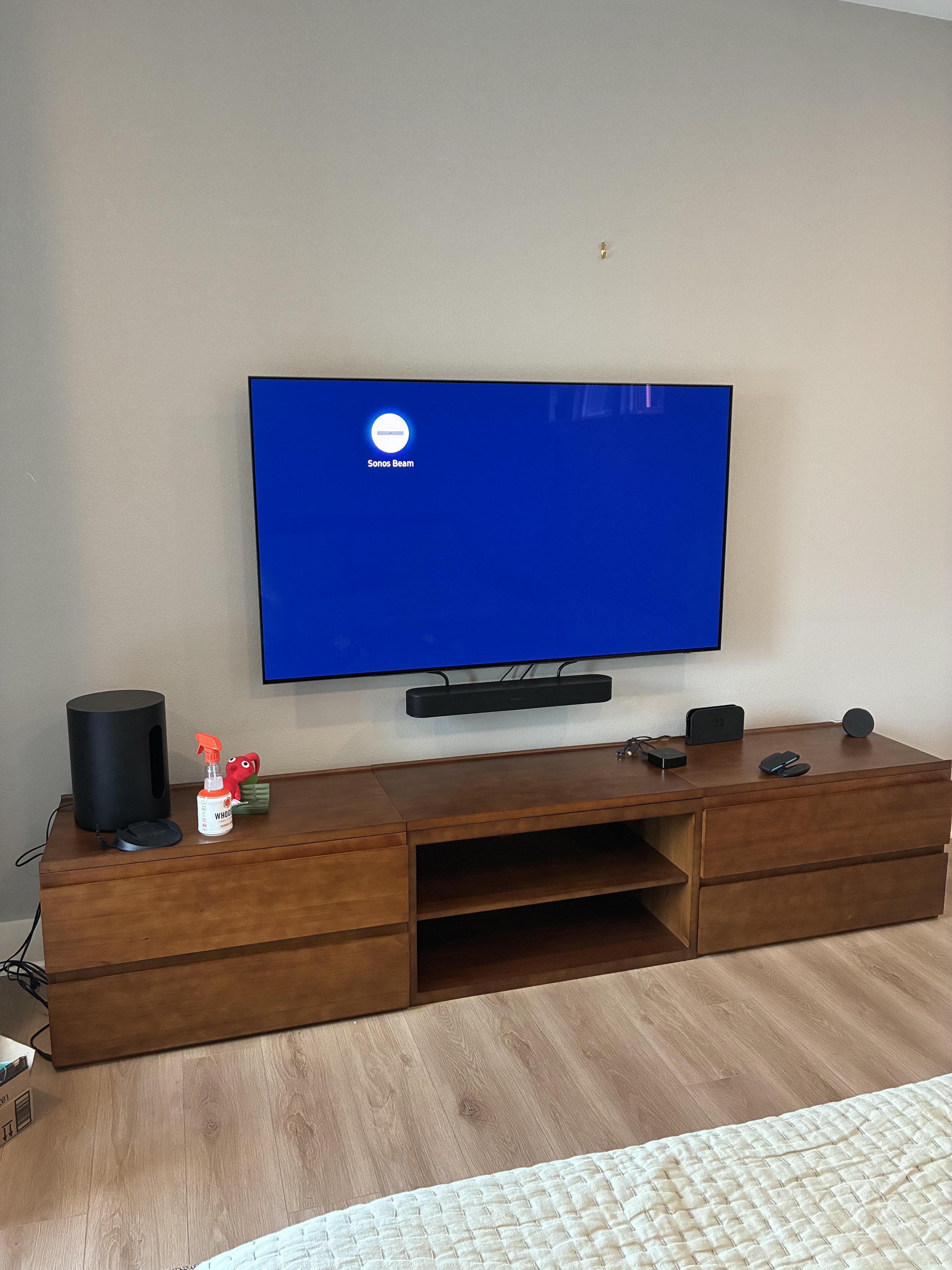Living room entertainment setup with a mounted flat-screen TV, Sony soundbar, a television remote, wireless speaker, router, and a few small decorative items on a wooden entertainment console.