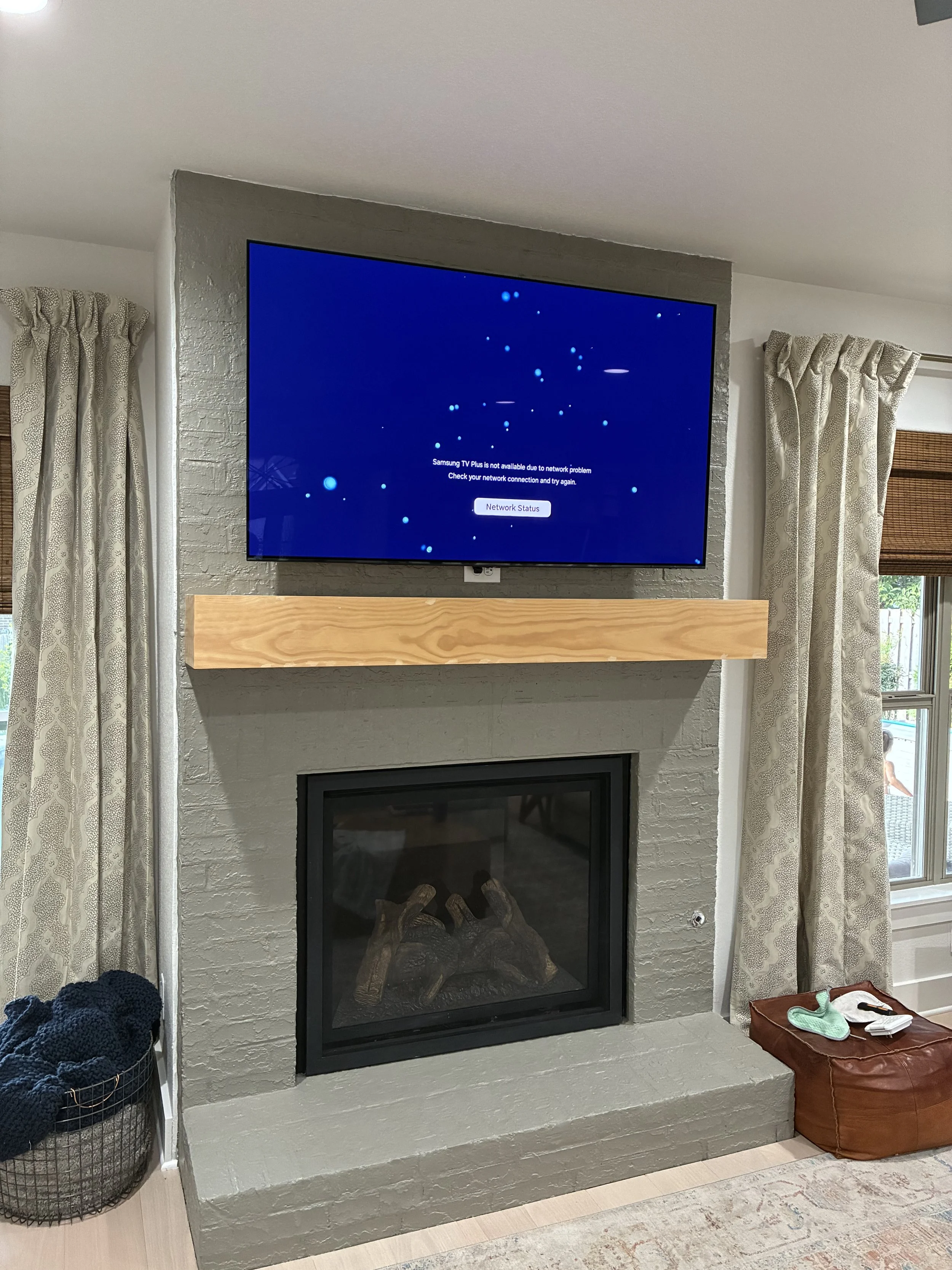 A living room with a mounted flat-screen TV displaying a network error message, a fireplace with decorative logs, and a wooden mantel. Curtains and a window are on either side of the fireplace, and a basket with a blanket is on the left, with a brown