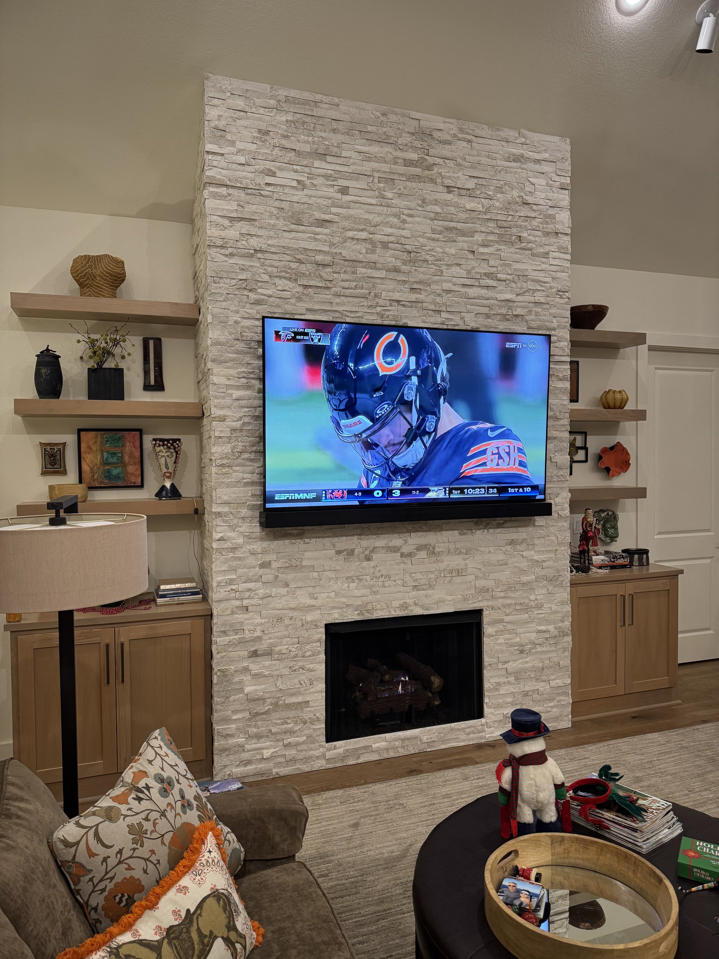 Living room with a wall-mounted flat-screen TV showing a football game, a fireplace below the TV, a sectional sofa with patterned pillows, a table with holiday decorations, and built-in shelves with decorative items.