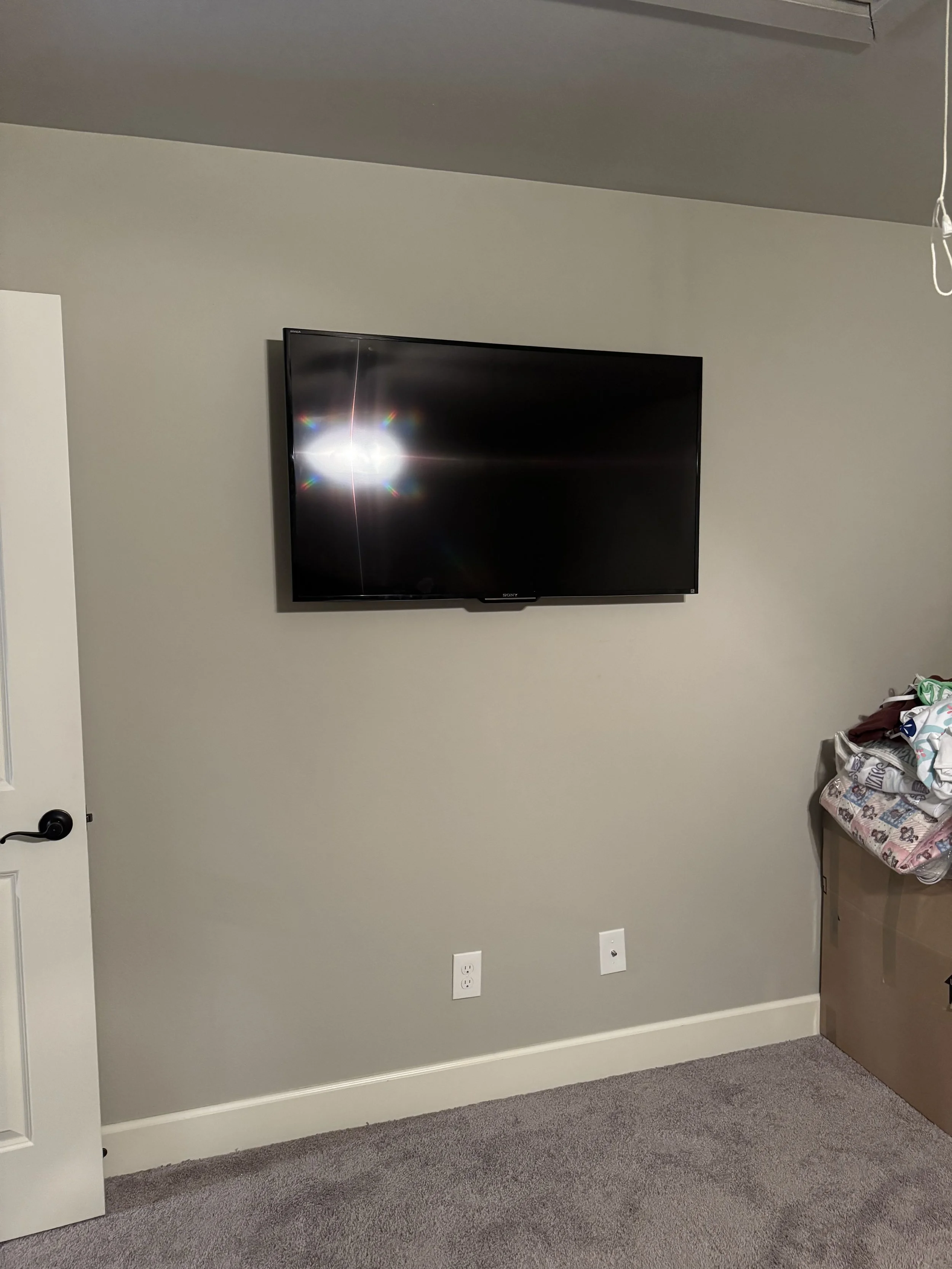 A living room wall with a mounted flat-screen TV, electrical outlets, and part of a door to the left. To the right, there is a cardboard box with a pile of folded and crumpled clothing or fabric.