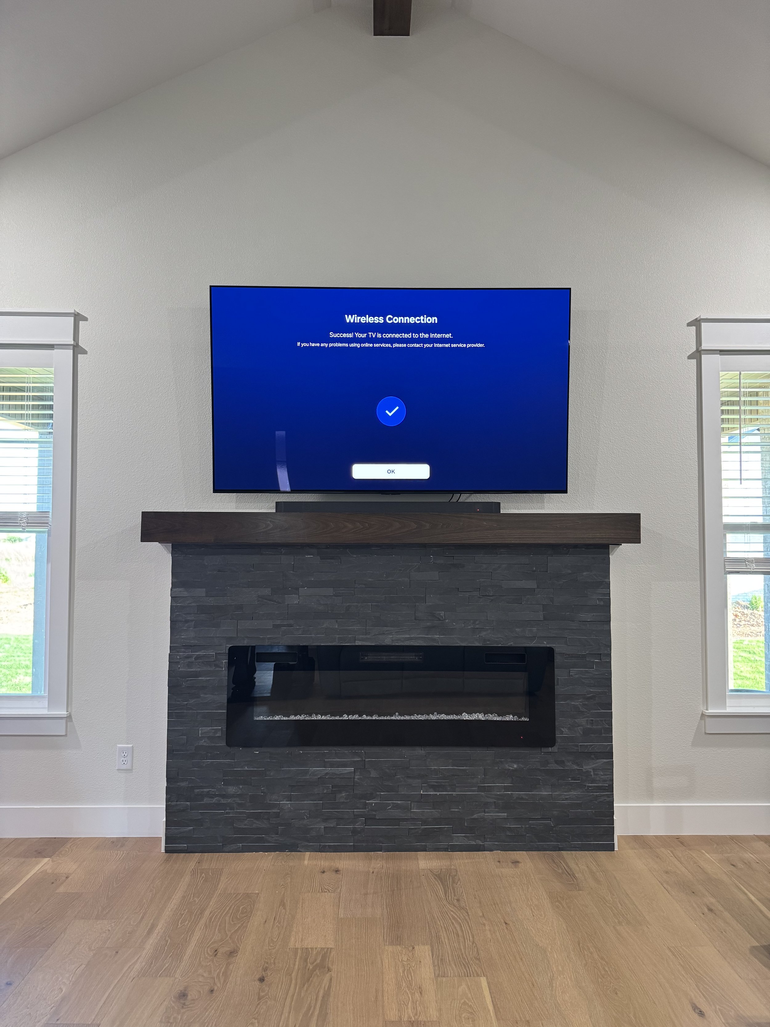 Living room with electric fireplace, mounted flat-screen TV displaying a successful wireless connection message, and two windows with white blinds on a beige wall.
