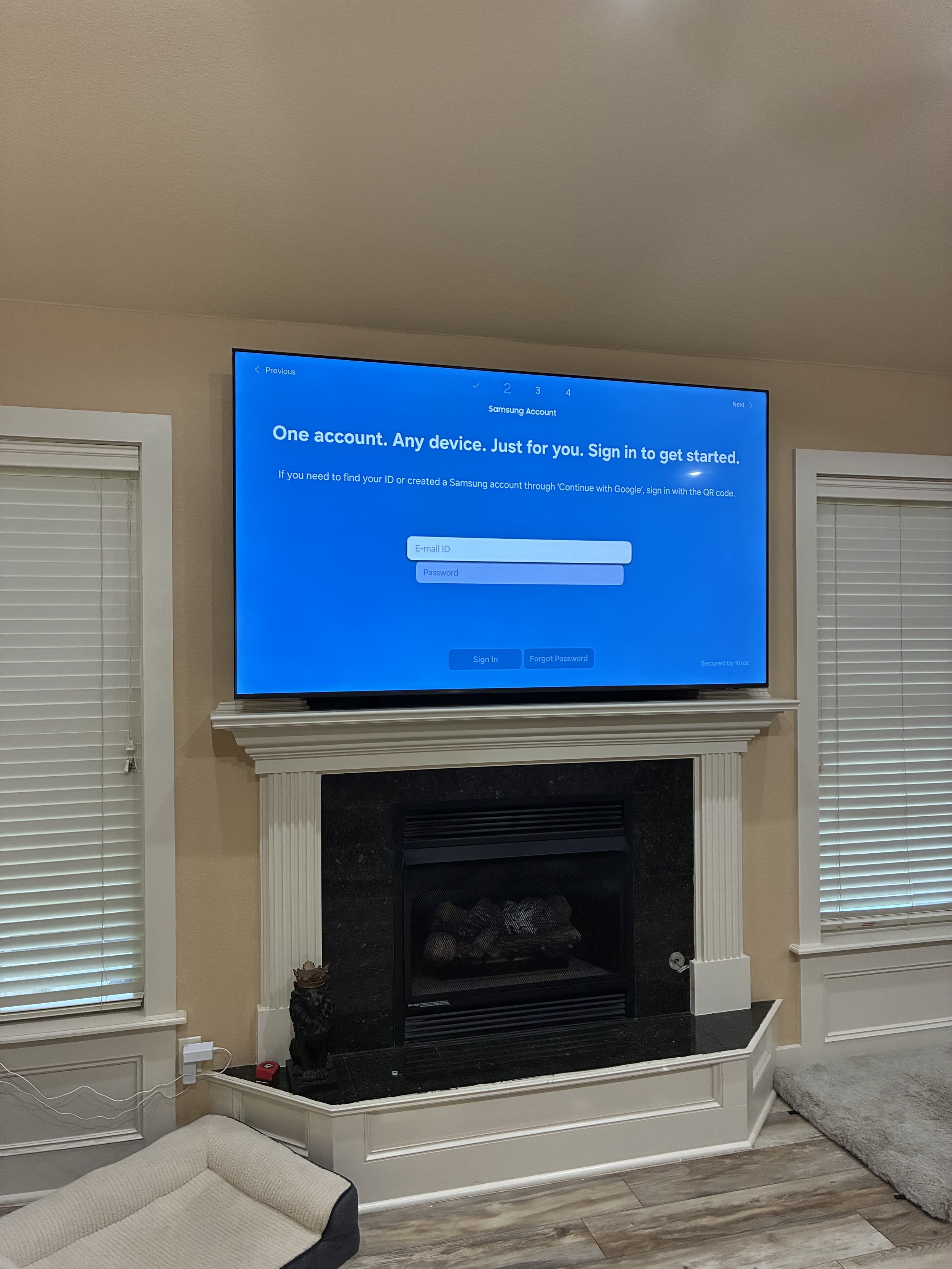 A living room with a mounted flat-screen TV displaying a Samsung account sign-in page above a fireplace with white molding and black hearth. To the left, there are two windows with closed white blinds. A small beige and black pet bed is on the floor 