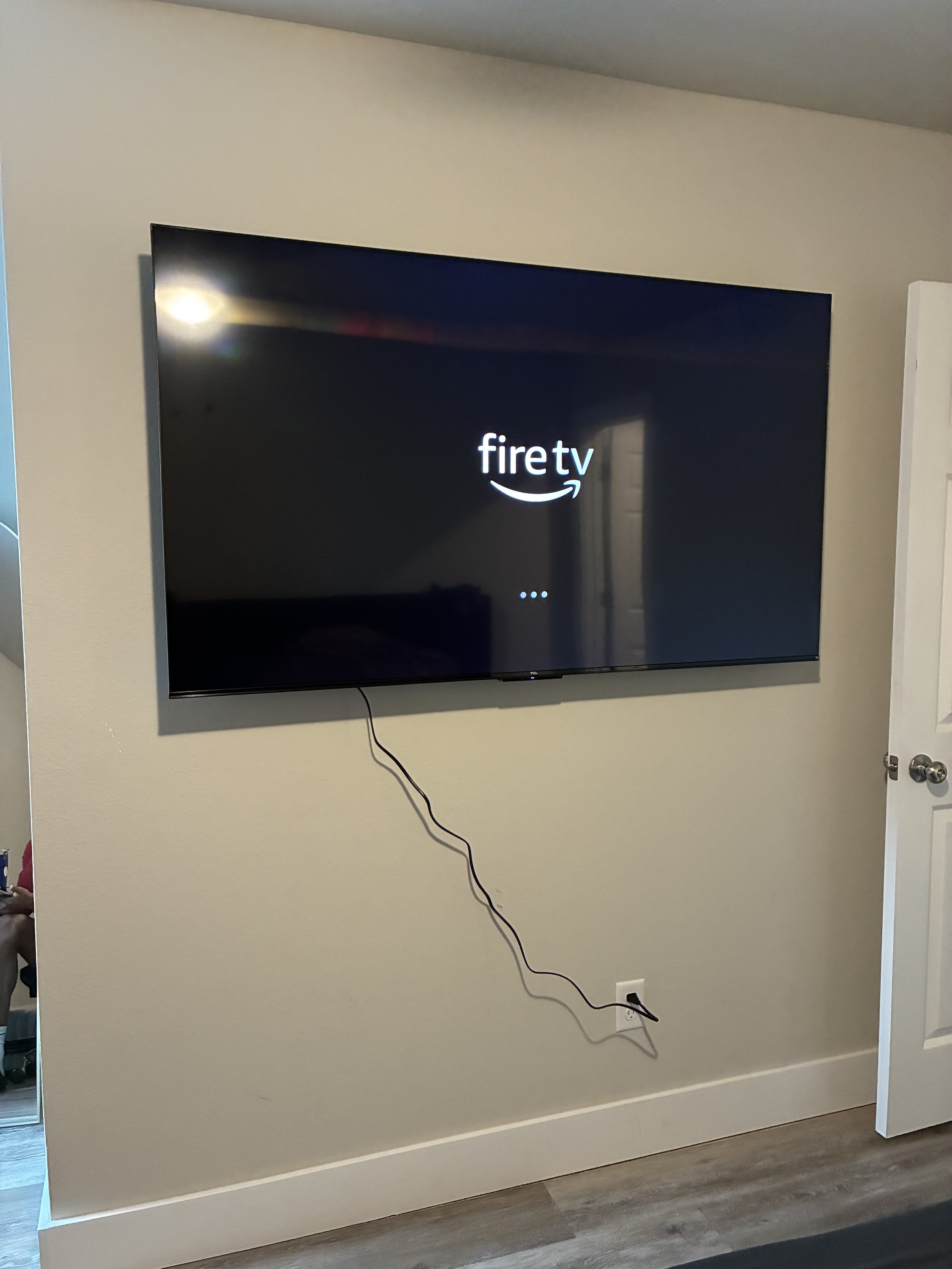 A flat screen fire TV mounted on a beige wall, displaying the 'fire tv' logo with loading dots, with a black power cord hanging down and plugged into an outlet.