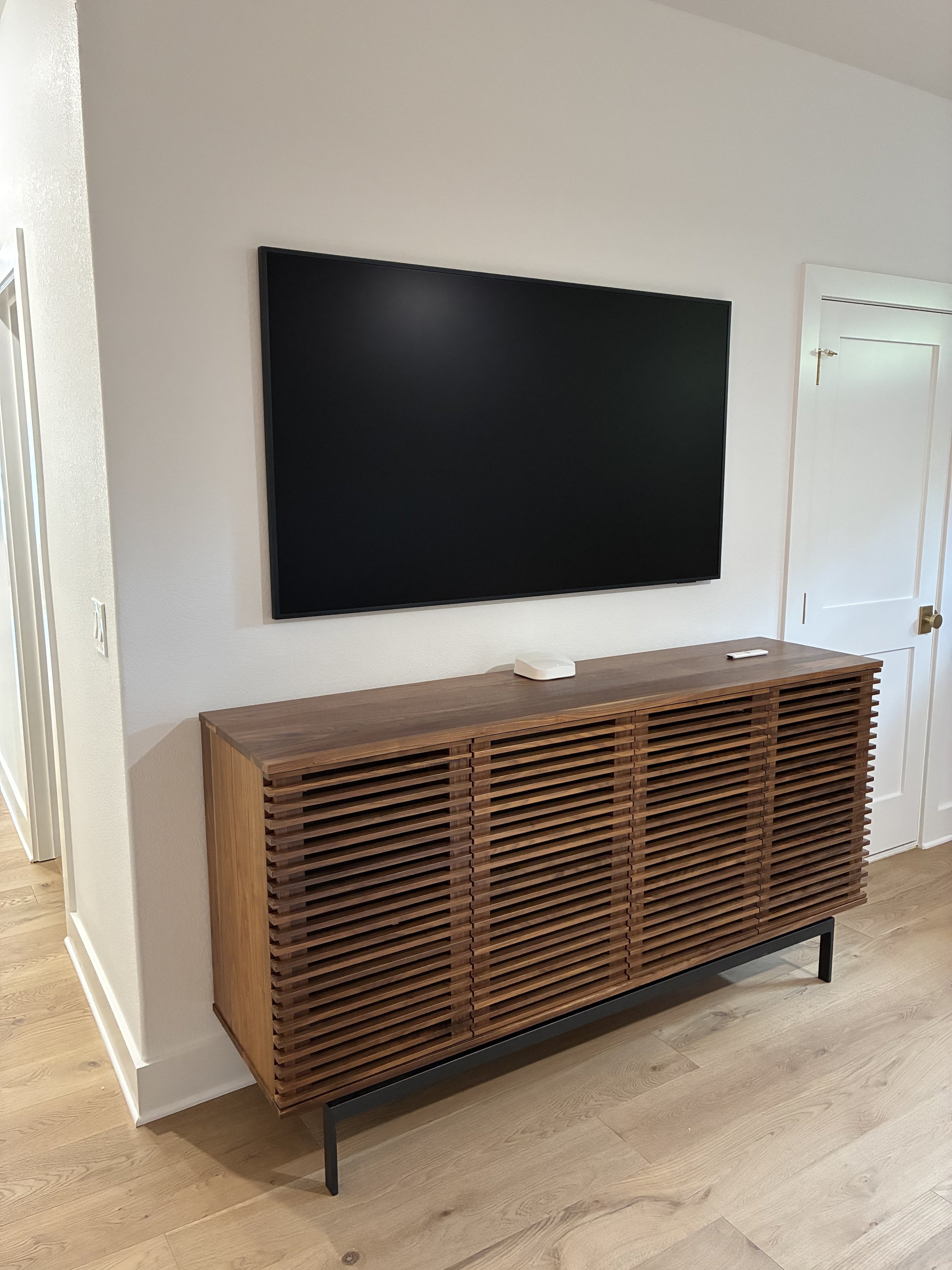 A flat-screen TV mounted on a white wall above a wooden credenza with a slatted front. On top of the credenza are a small white device and a remote control. The floor is light-colored hardwood.