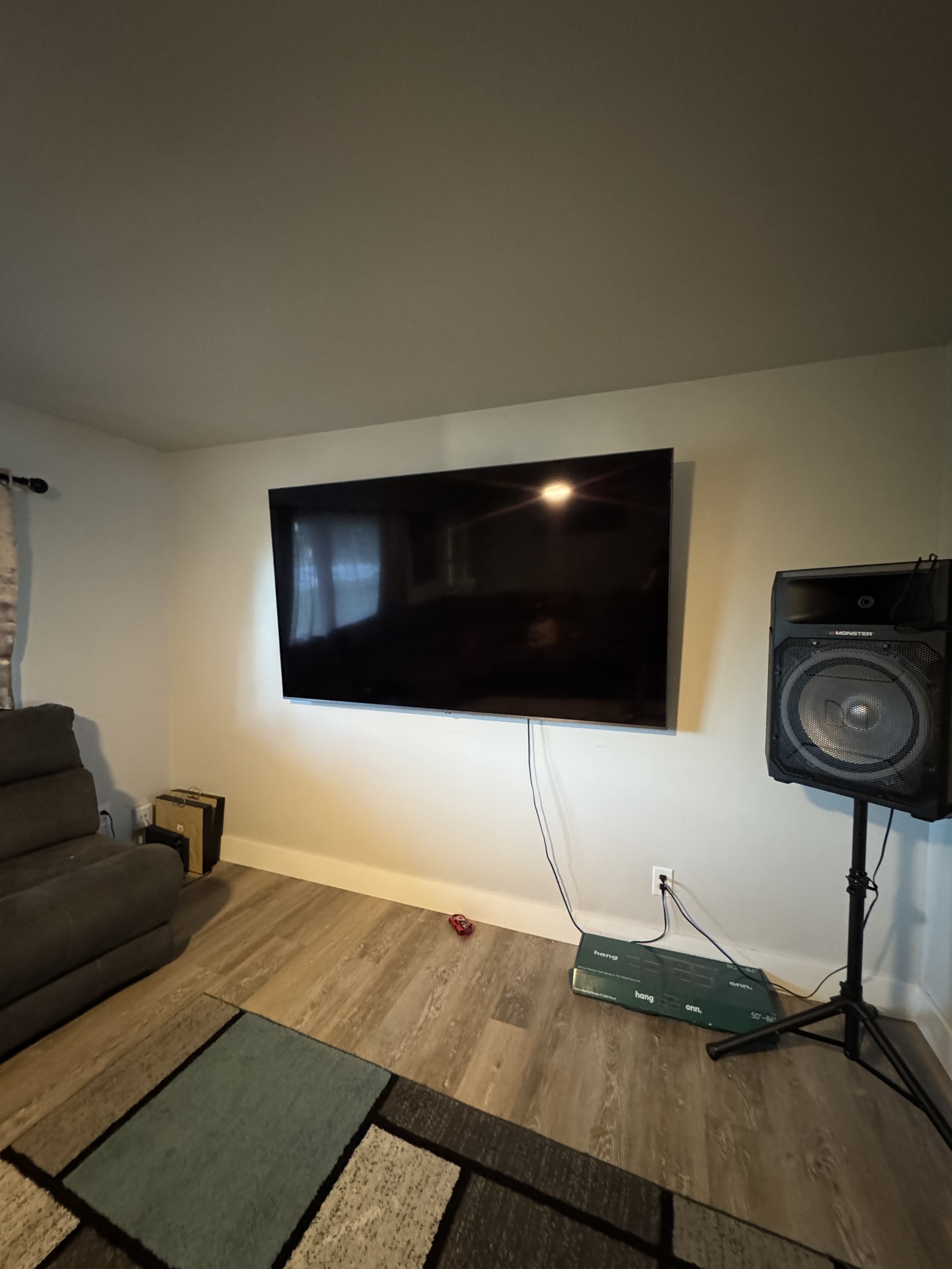 Living room with a large wall-mounted flat-screen TV, a speaker on a stand, a patterned rug, a brown couch, a window with curtains, and some electronic devices on the floor.