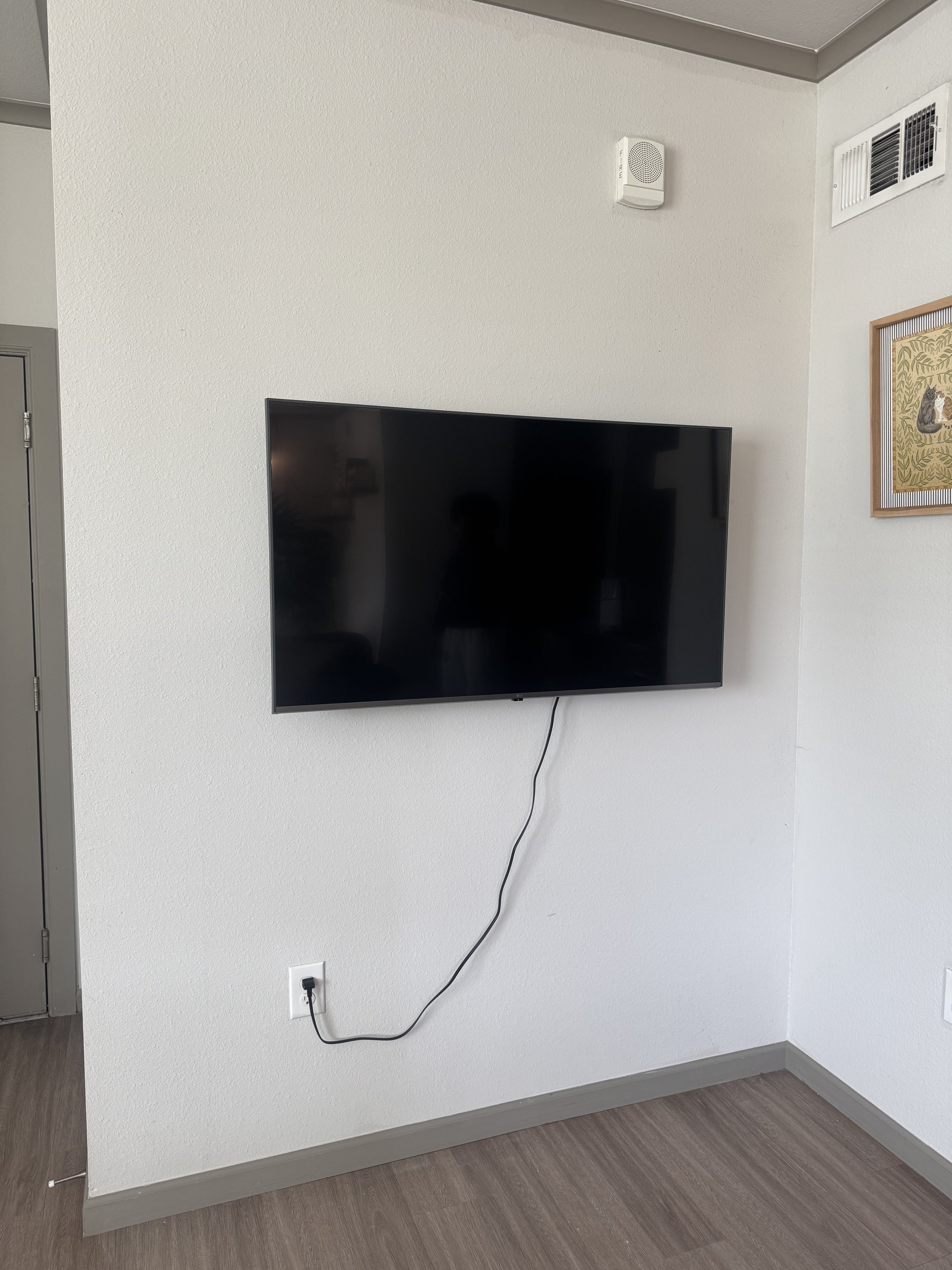 A flat-screen TV mounted on a white wall with a black cable plugged into an outlet below, and a framed picture on the right side.