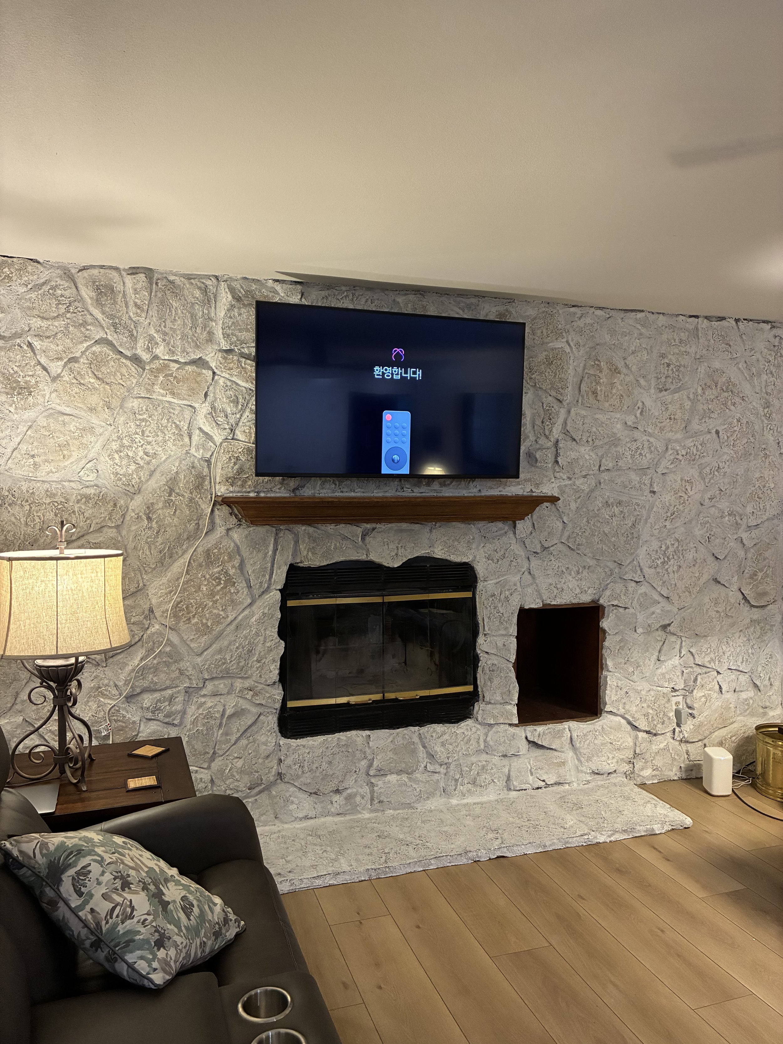 Living room with a stone fireplace, a flat-screen TV mounted above the mantel with the message "Welcome" in Korean, a side table with a lamp, a couch with a floral pillow, and corner decorations including a white speaker and a gold container.