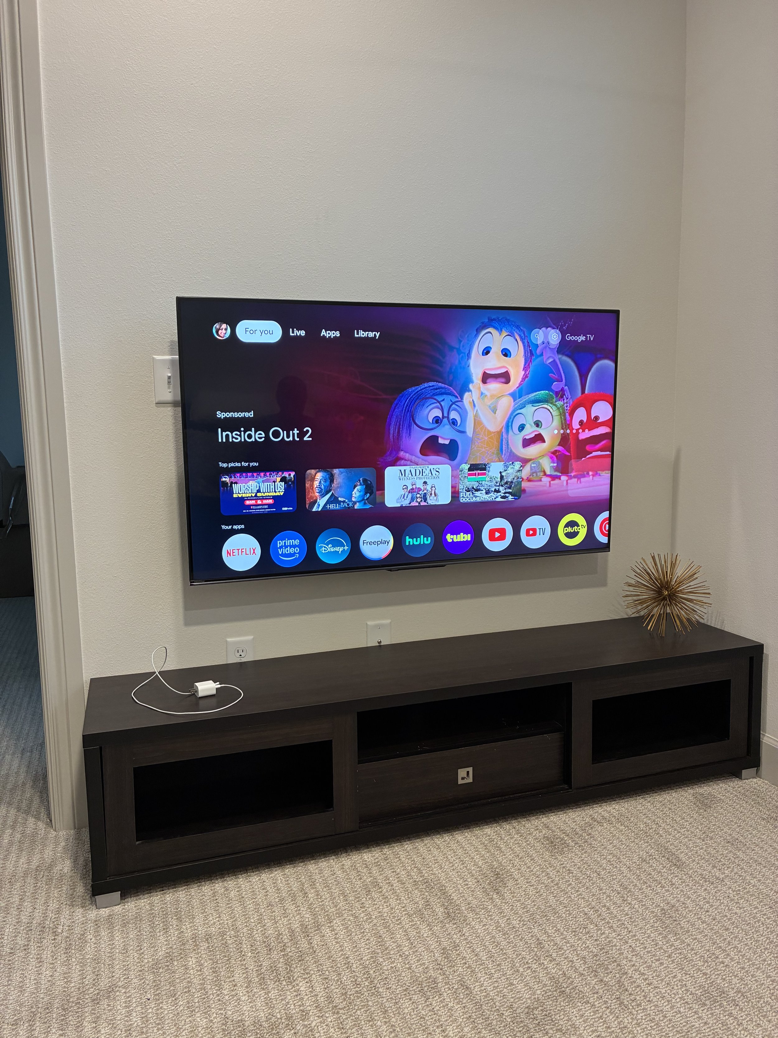 A wall-mounted flat-screen TV displaying the streaming app menu with characters from Inside Out 2, sitting on a dark wooden TV stand with open shelves, beside a decorative golden spiked sphere on the right.