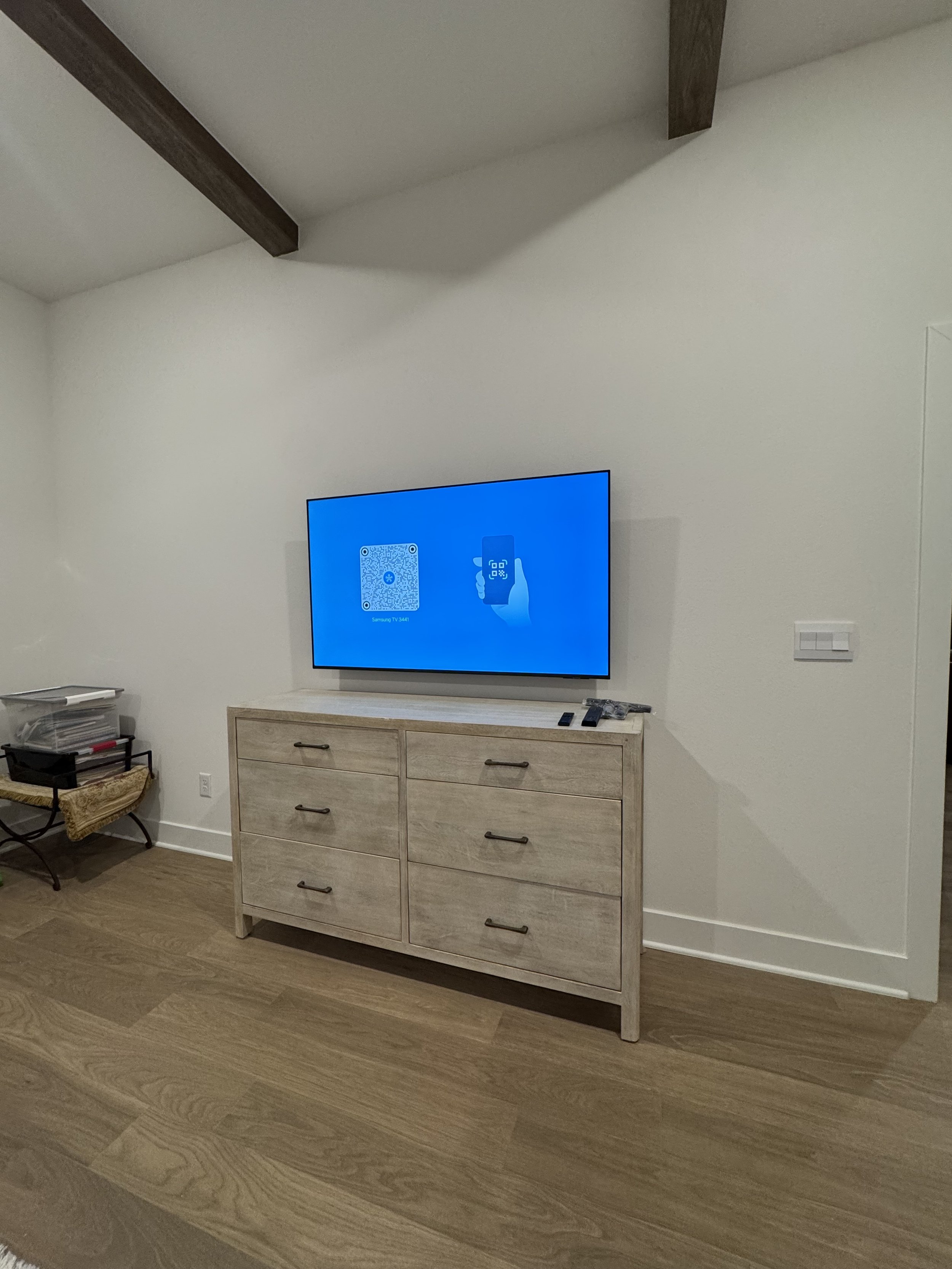 Modern living room corner with wall-mounted flat-screen TV displaying a QR code and an illustration of a hand holding a phone, a light-colored wooden dresser underneath, a small black side table with books and other items, and wood flooring.