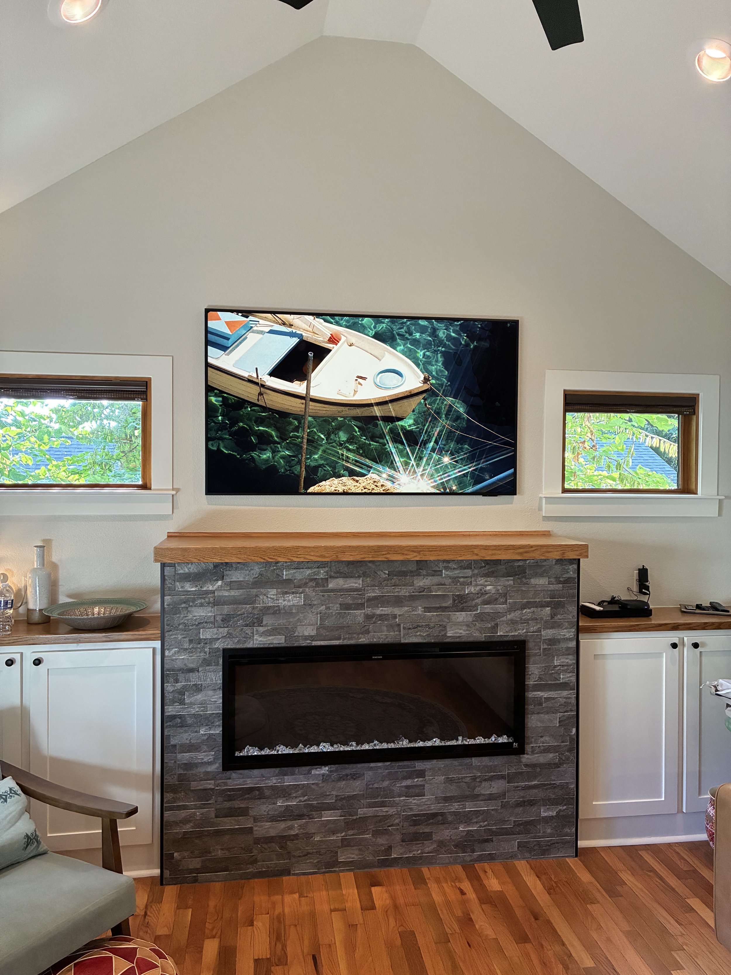 Living room fireplace with a flat-screen TV mounted above it, showing a boat on water. Two small windows with green foliage outside, white cabinets on each side of the fireplace, and wooden flooring.