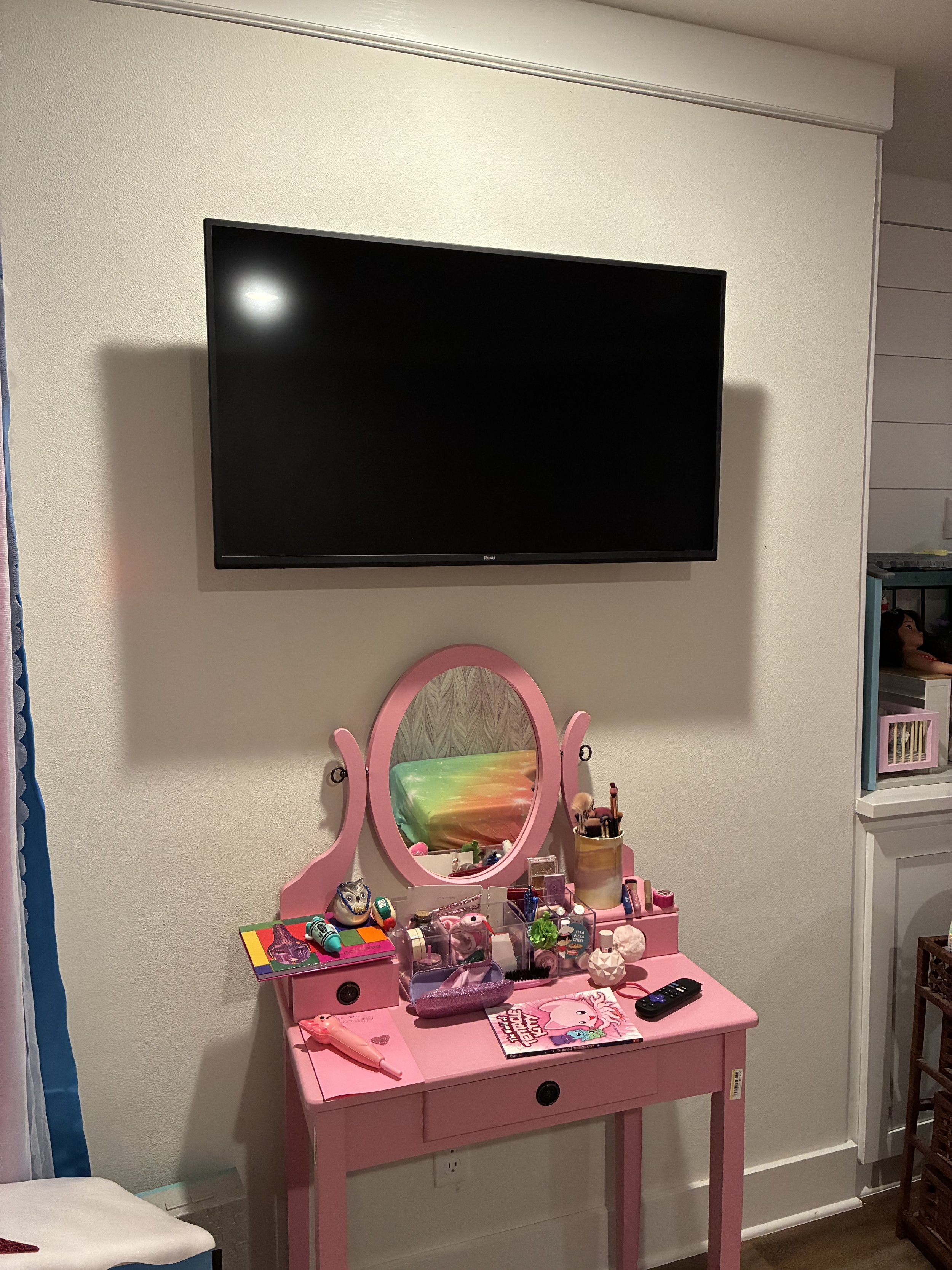 Pink children's vanity table with a mirror, items like toys, makeup, and books, situated below a wall-mounted flat-screen TV in a room.