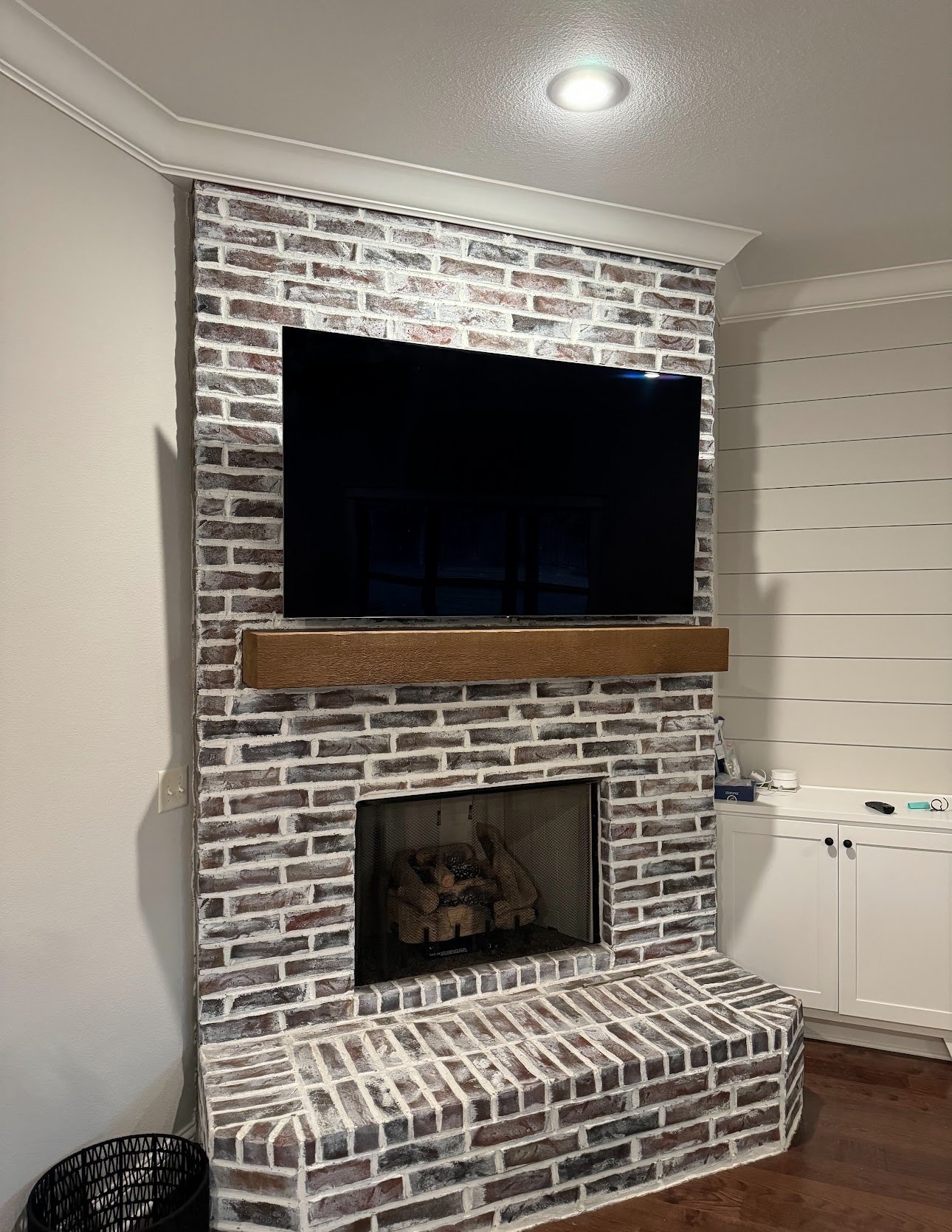 A brick fireplace with a flat-screen TV mounted above it and a wooden shelf below the TV in a living room.