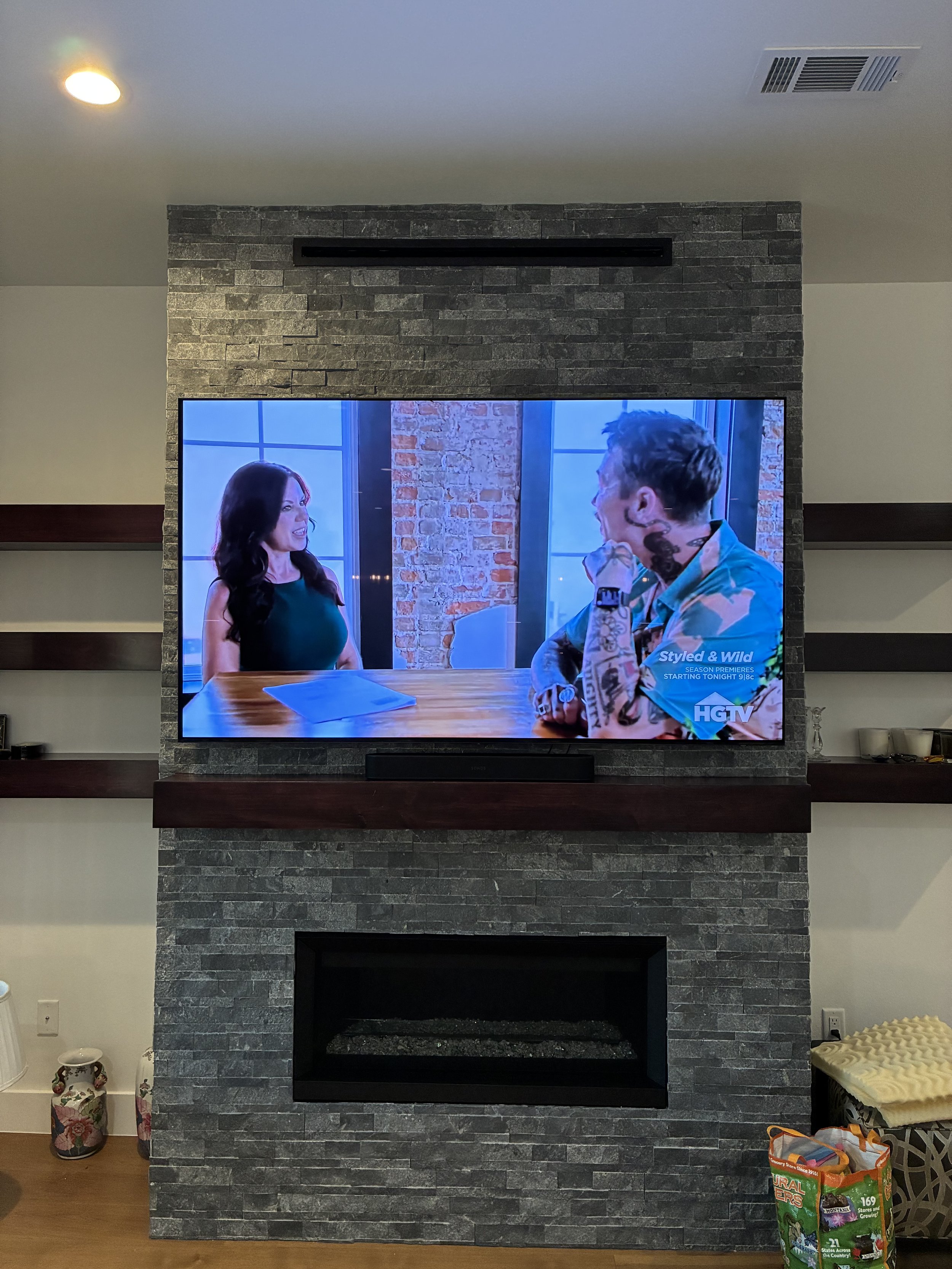 Living room with a flat-screen TV mounted above a modern gray brick fireplace, with shelves on either side of the fireplace, and various decor on the shelves and floor.
