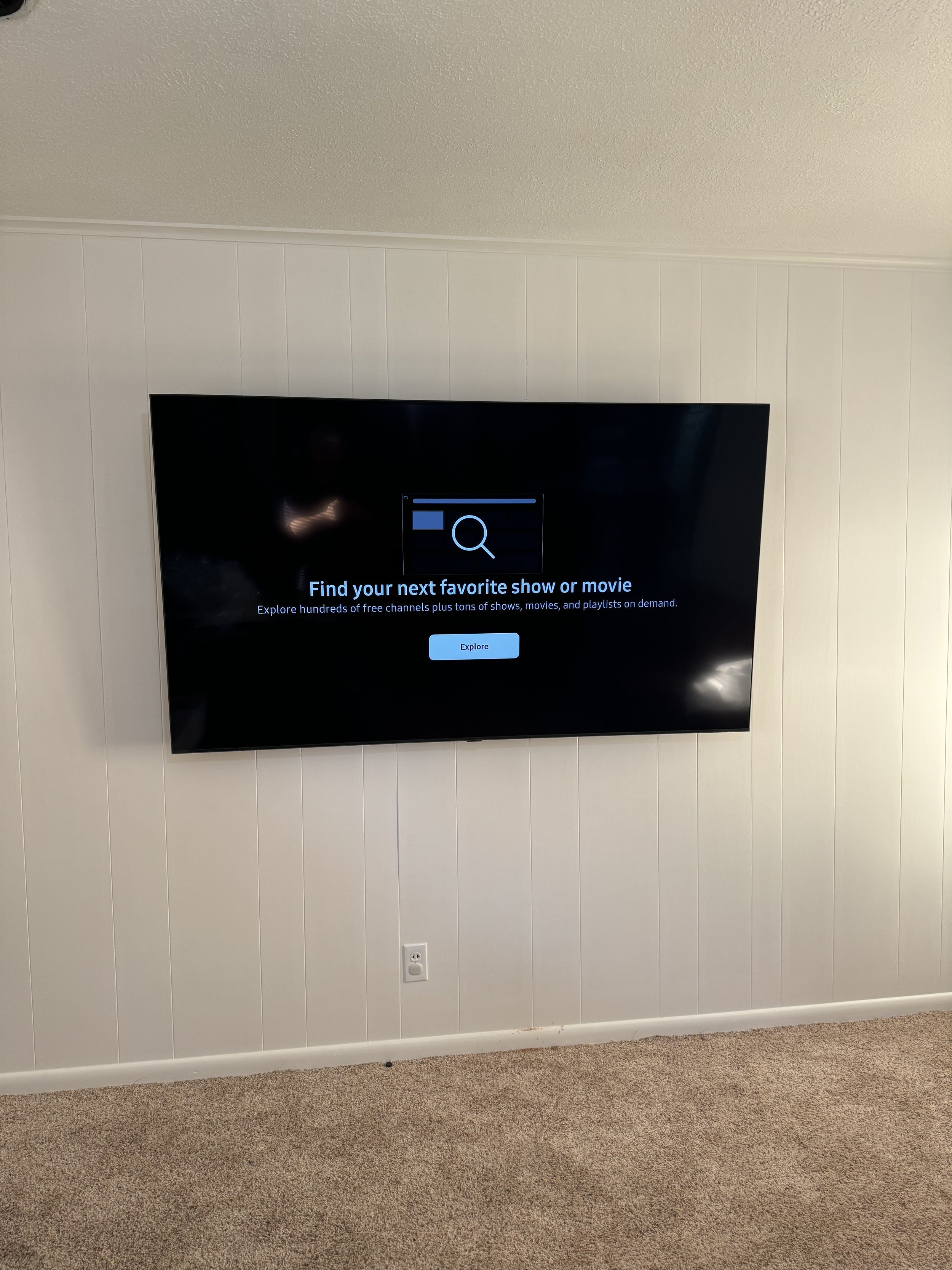 Wall-mounted television displaying a screen with a search magnifying glass icon, a message to find your favorite show or movie, and a blue explore button, in a room with beige carpet and white paneled walls.