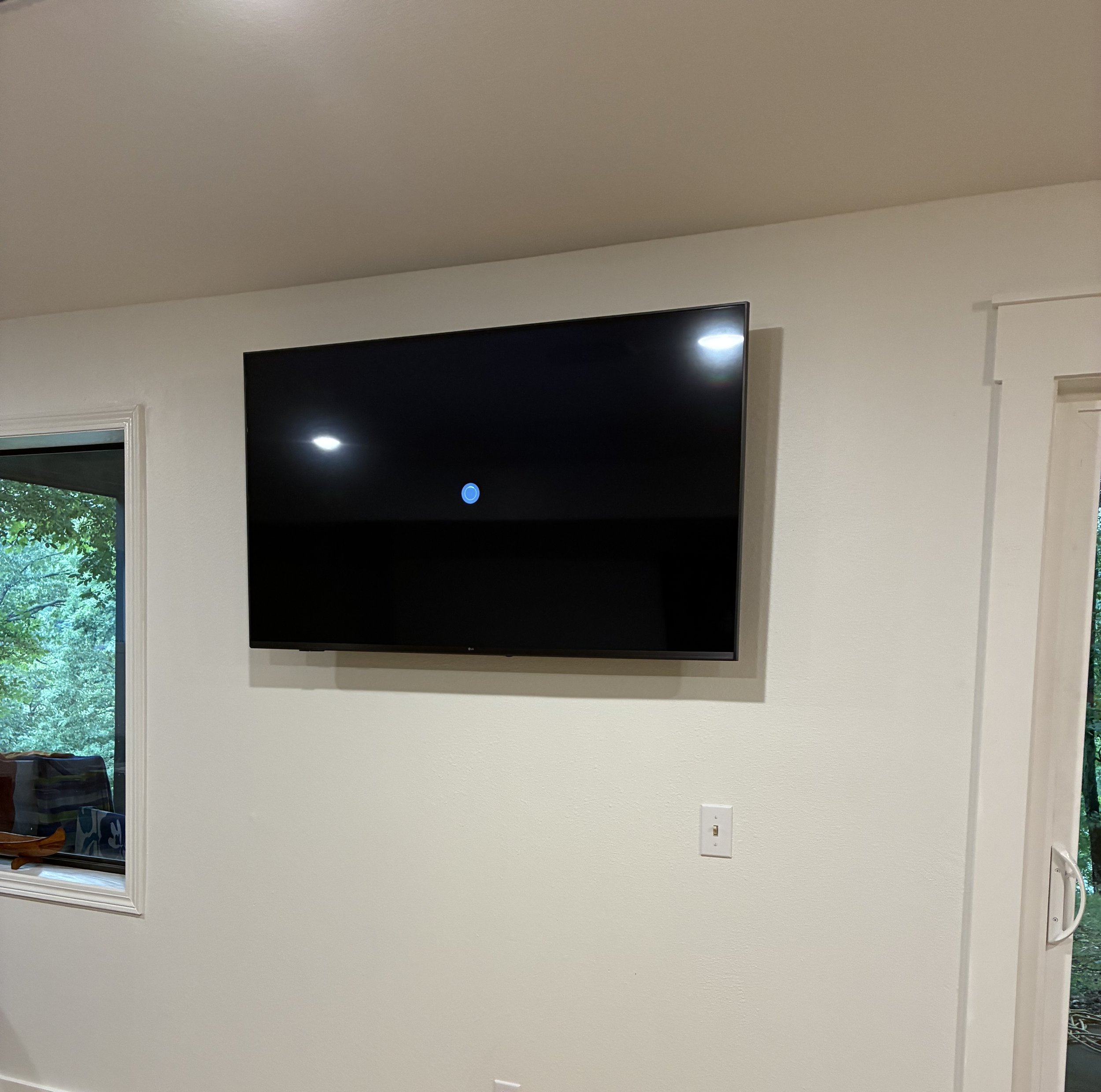 A flat-screen TV mounted on a white wall in a room with a window showing greenery outside.