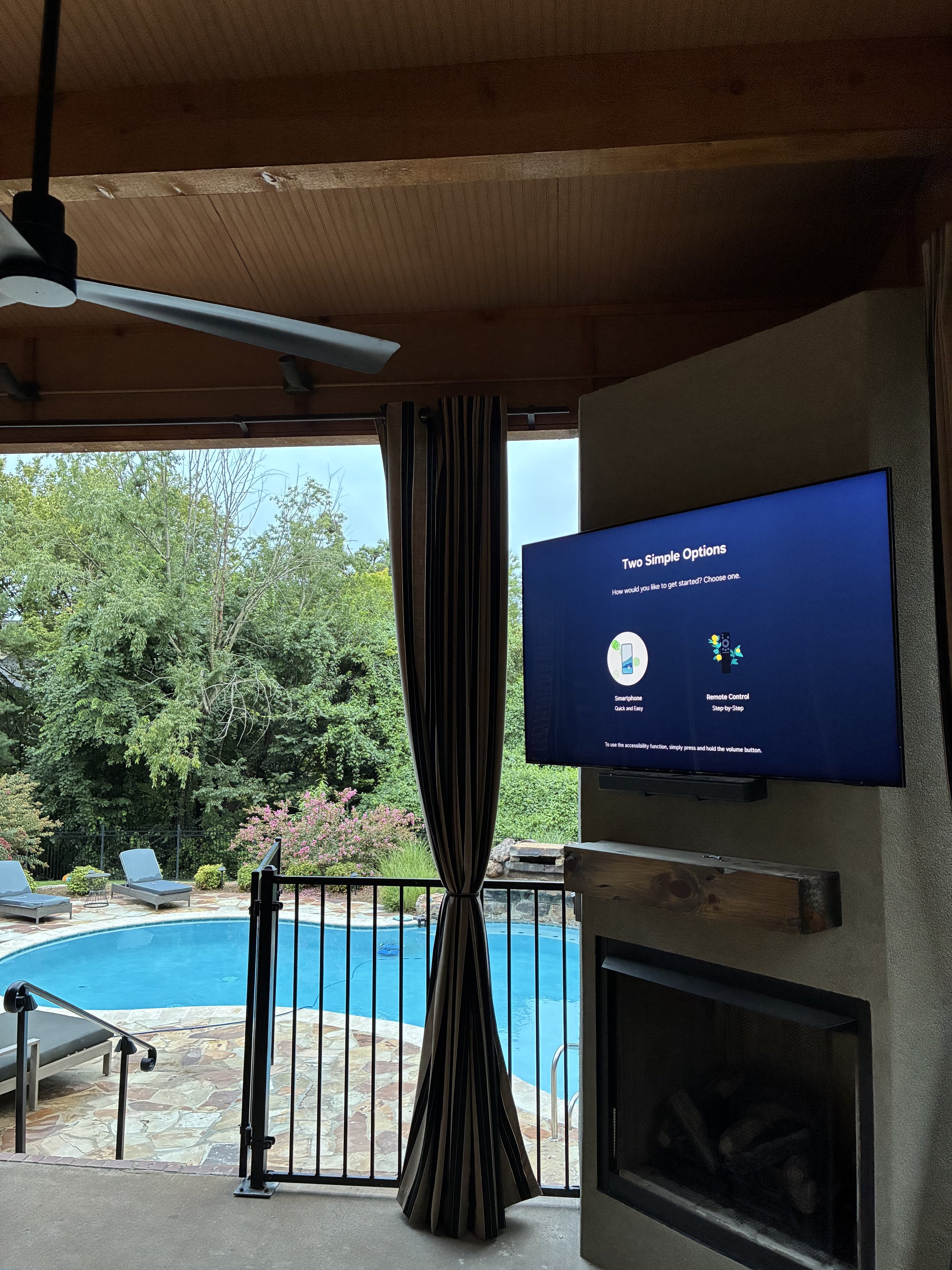 Living room with a television displaying two options, overlooking a backyard pool with lounge chairs and green trees.