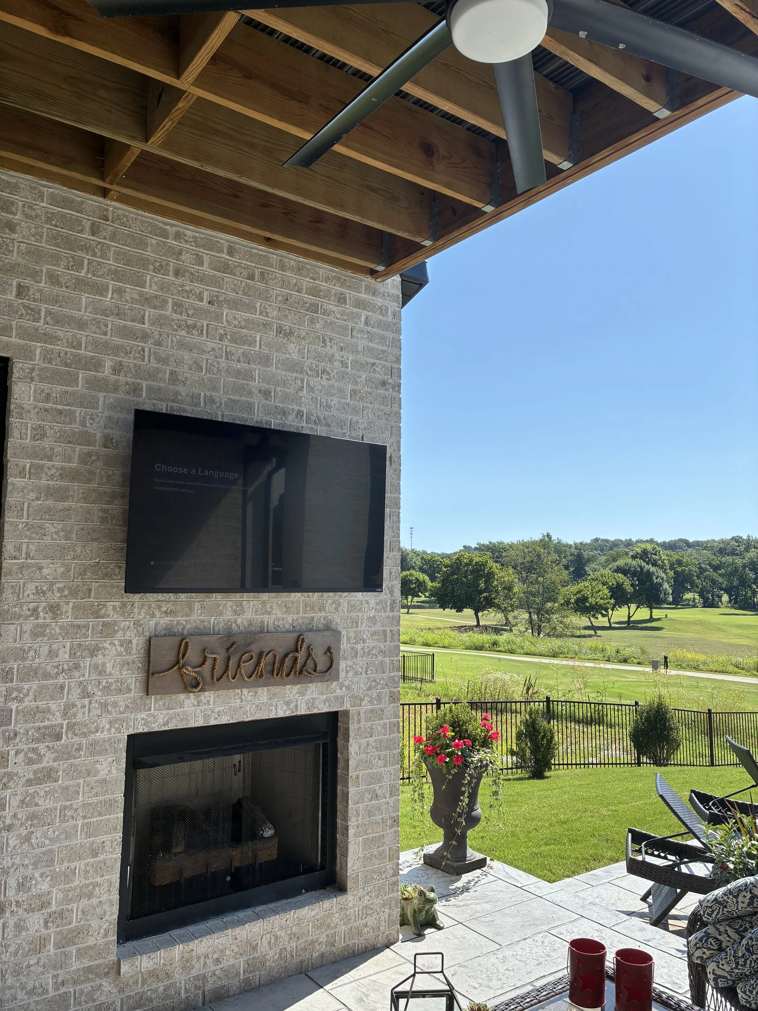 Outdoor patio with a stone fireplace, TV, a wooden sign reading 'friends,' potted flowers, and chairs overlooking a grassy field with trees under a clear blue sky.