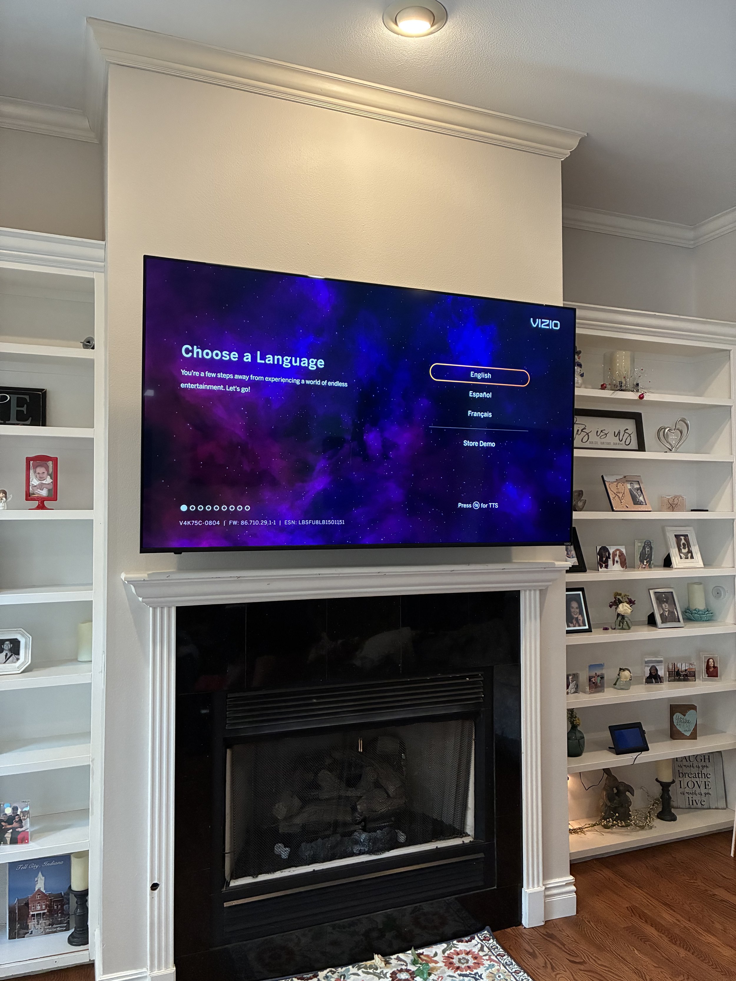 A living room with a wall-mounted television displaying a language selection screen, above a black fireplace with a white mantel, flanked by white bookshelves filled with framed photos and decorative items, and a floral area rug on a hardwood floor.