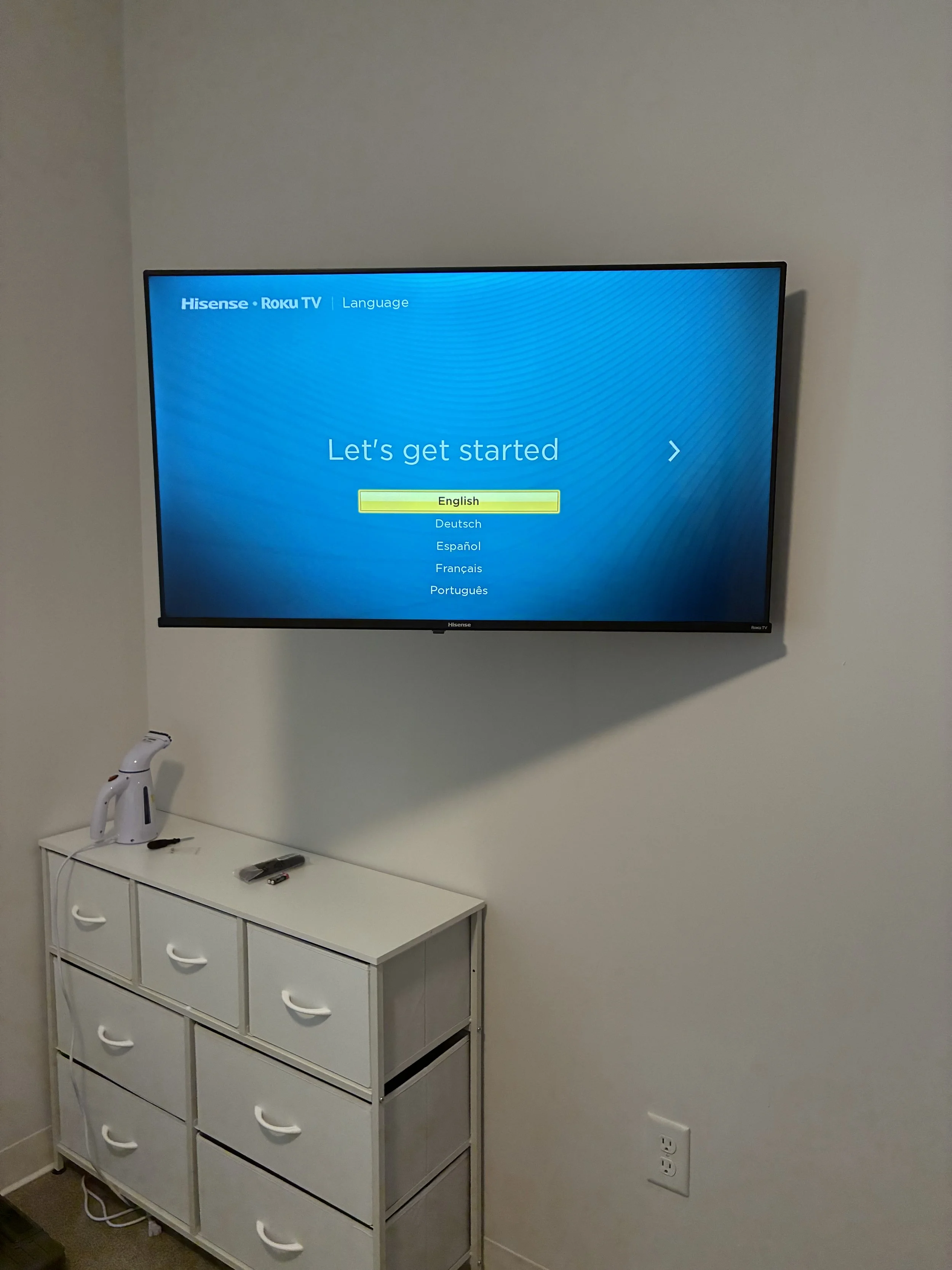 A wall-mounted flat-screen TV displaying a language selection screen, with options including English, Deutsch, Español, Français, and Português. Below the TV, a white cabinet with drawers holds a handheld device, a TV remote, and a small electronic d