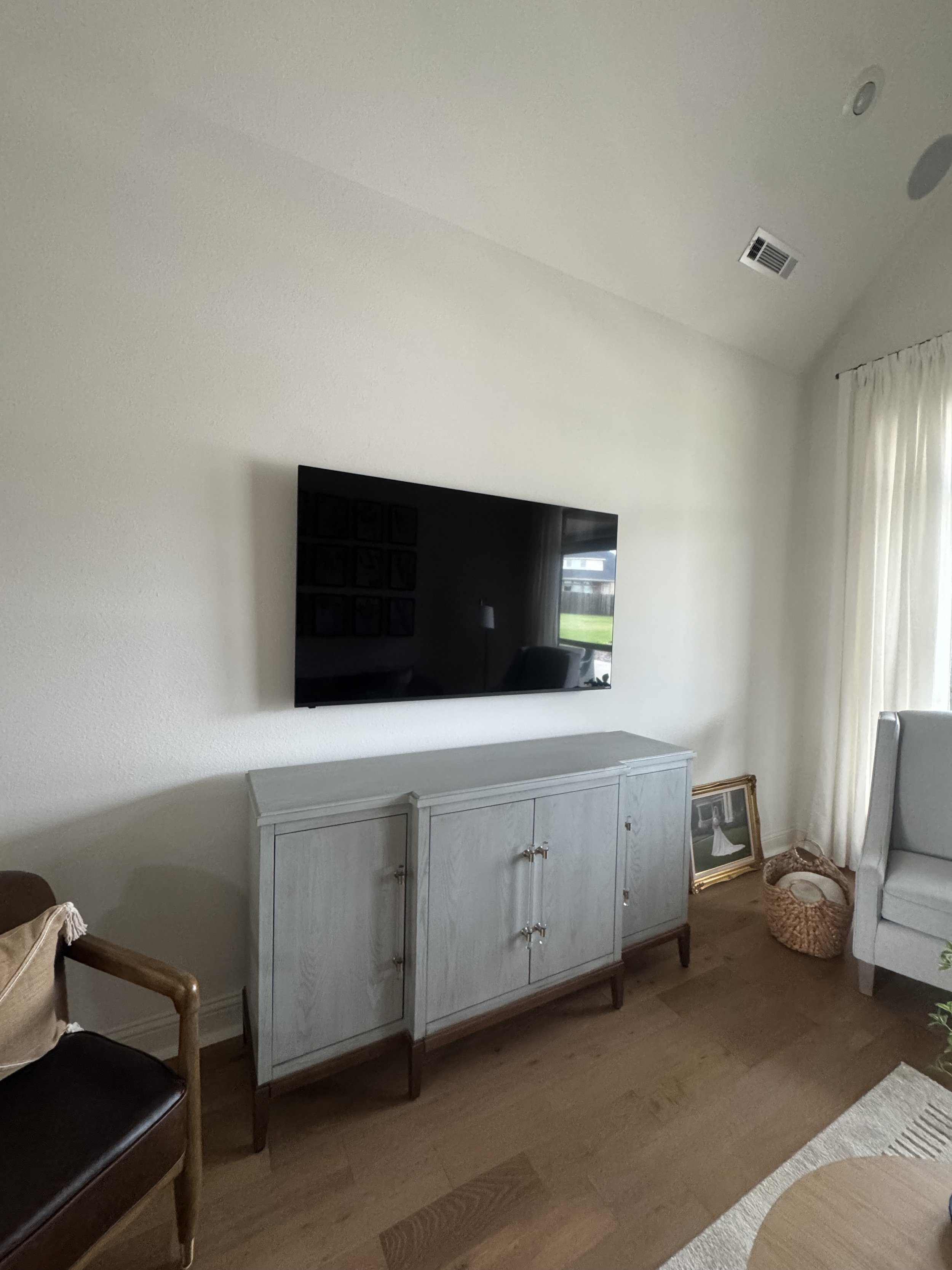 Living room with a mounted flat-screen TV on a white wall, a light-colored wooden cabinet below it, a framed picture leaning against the wall, a basket, and a light-colored armchair on the right. Curtains cover a window to the right.