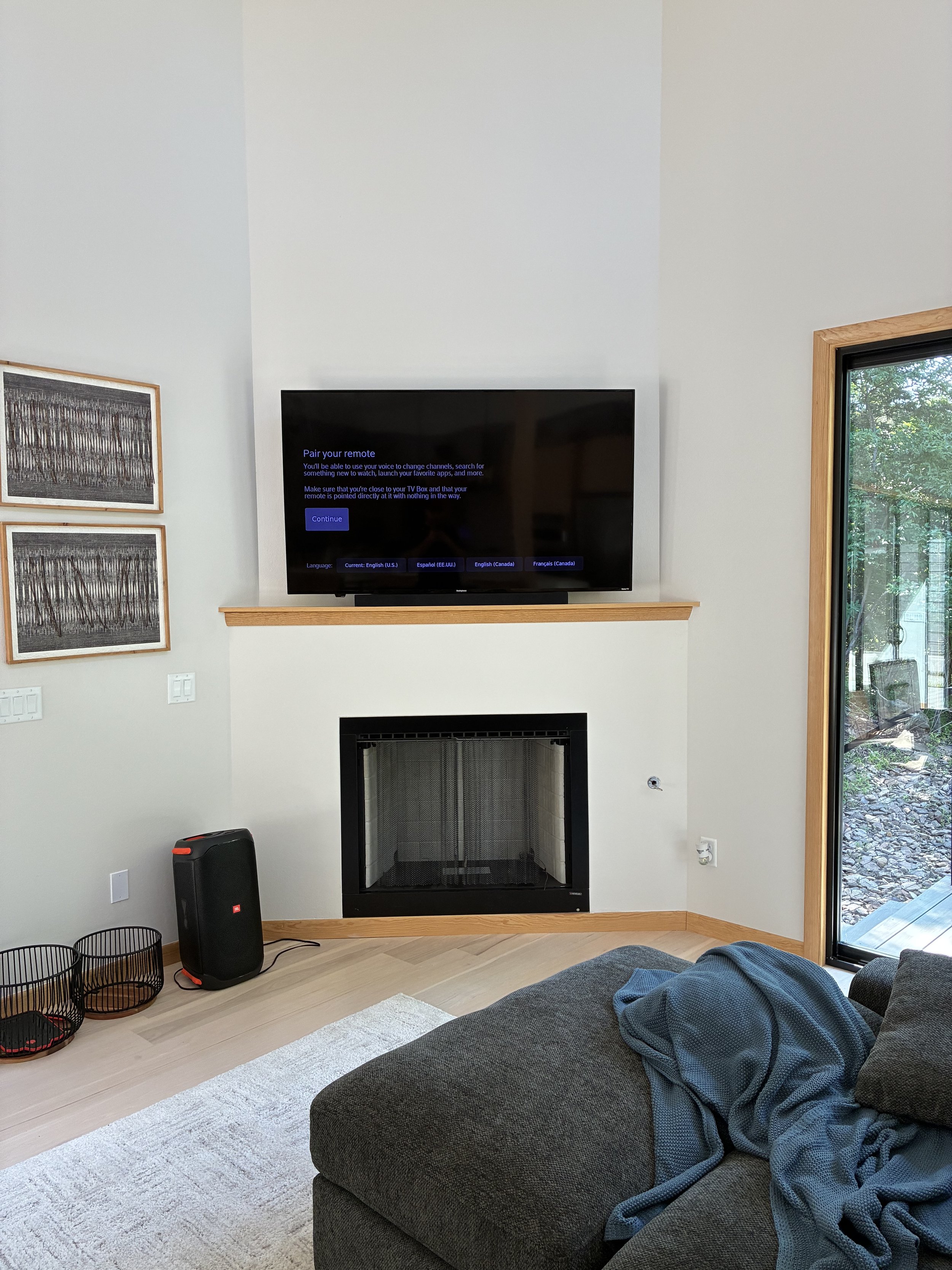 Living room with a wall-mounted TV displaying a pairing instruction, a fireplace beneath it, a gray sofa with a blanket, a sliding glass door leading outside, and decorative artwork on the wall.
