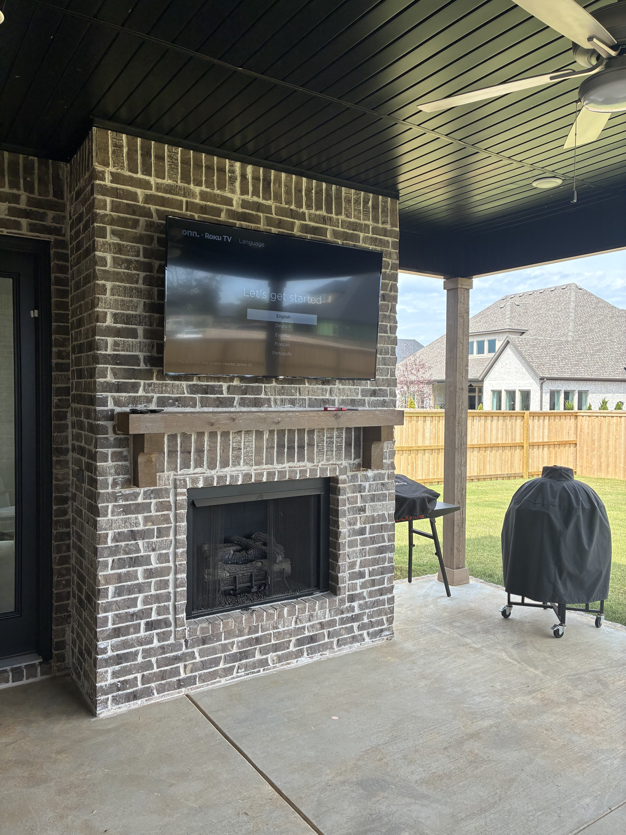 Outdoor covered patio with brick fireplace, mounted flat-screen TV, and a barbecue grill covered with a black cover. The area overlooks a backyard with a grass lawn and neighboring houses.