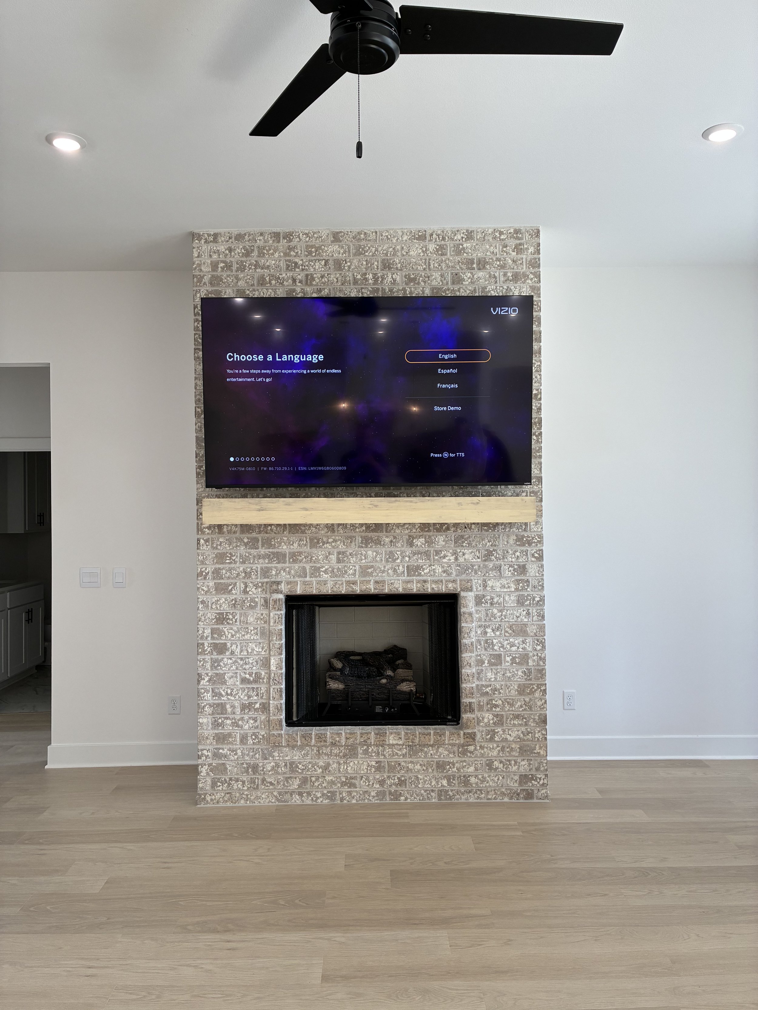 Living room with a brick fireplace and a mounted television screen above it, displaying a language selection menu. The room has a ceiling fan and recessed lighting, with light-colored wooden flooring and plain white walls.