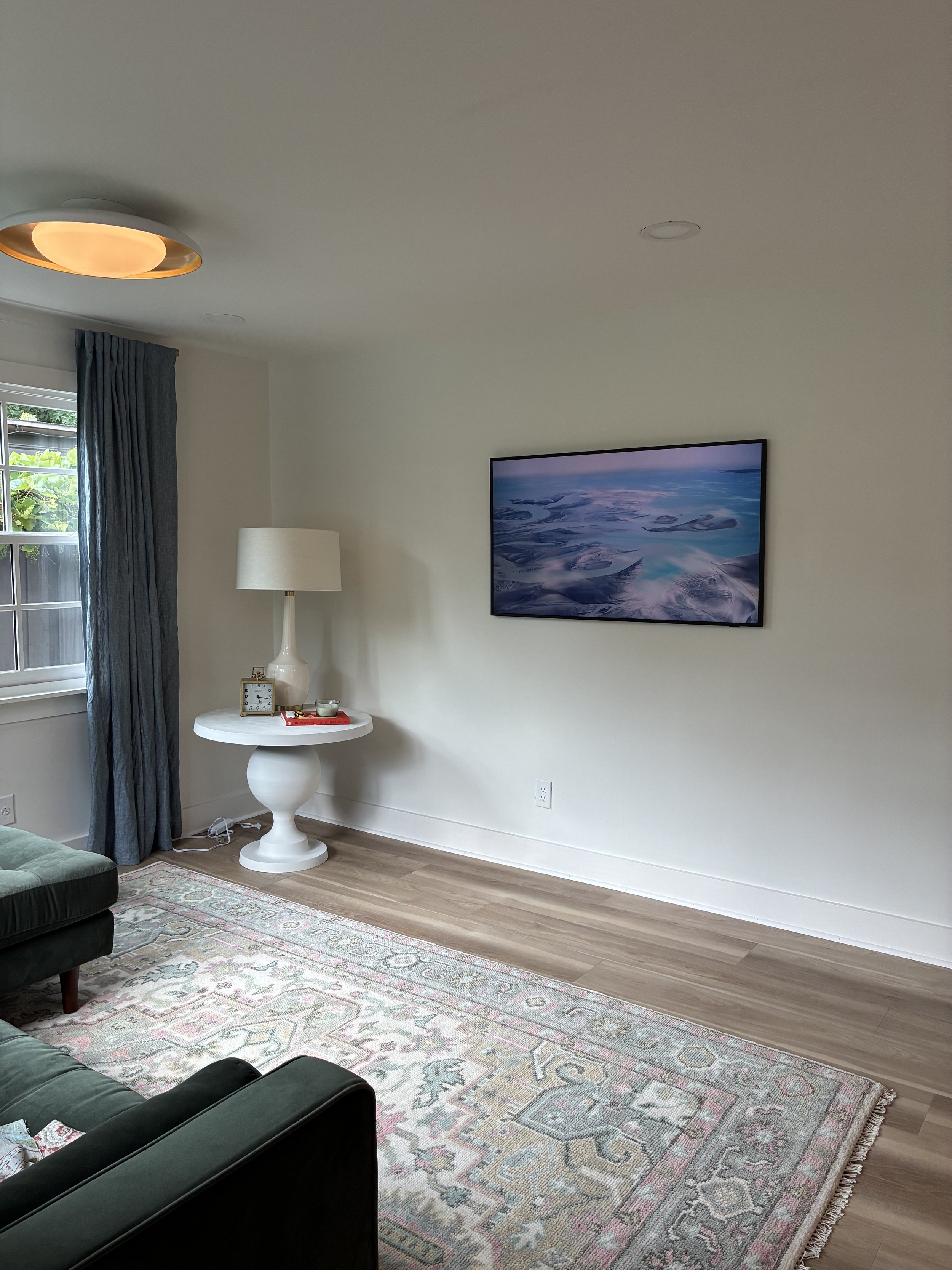 Living room with a window, dark curtains, a white table with a lamp, clock, and decor, a wall-mounted TV displaying a scenic landscape, hardwood flooring, a decorative rug, and part of a green sofa.