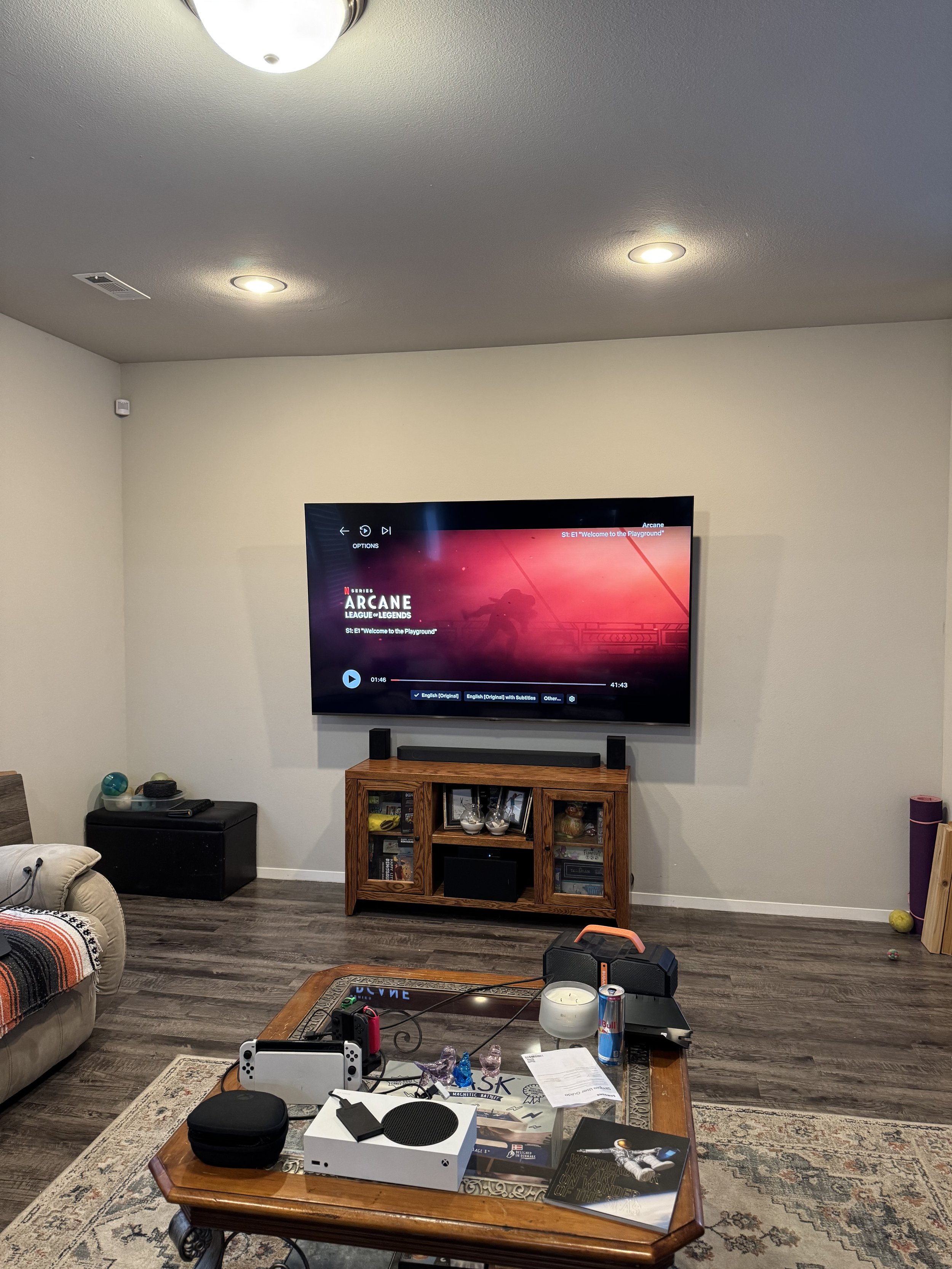 Living room with a wall-mounted TV displaying the game series 'Arcane,' a wooden entertainment center, a beige couch, a wooden coffee table with various items including a Nintendo Switch, a can of Red Bull, a candle, papers, and a book, a black speak