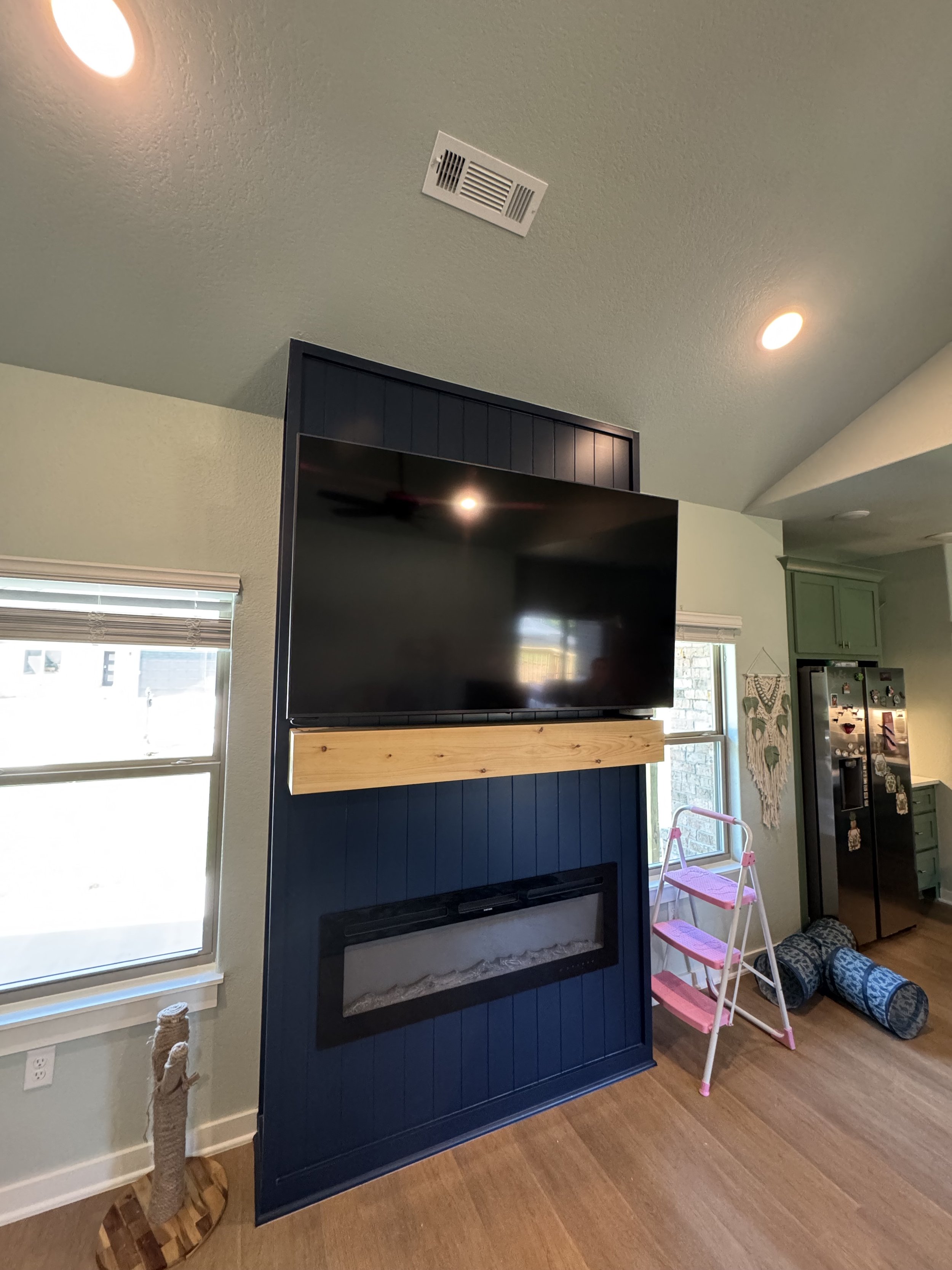 Living room with a large flat-screen TV mounted above a modern electric fireplace on a navy blue wall, with two windows on either side, a pink step ladder, and a refrigerator in the background.