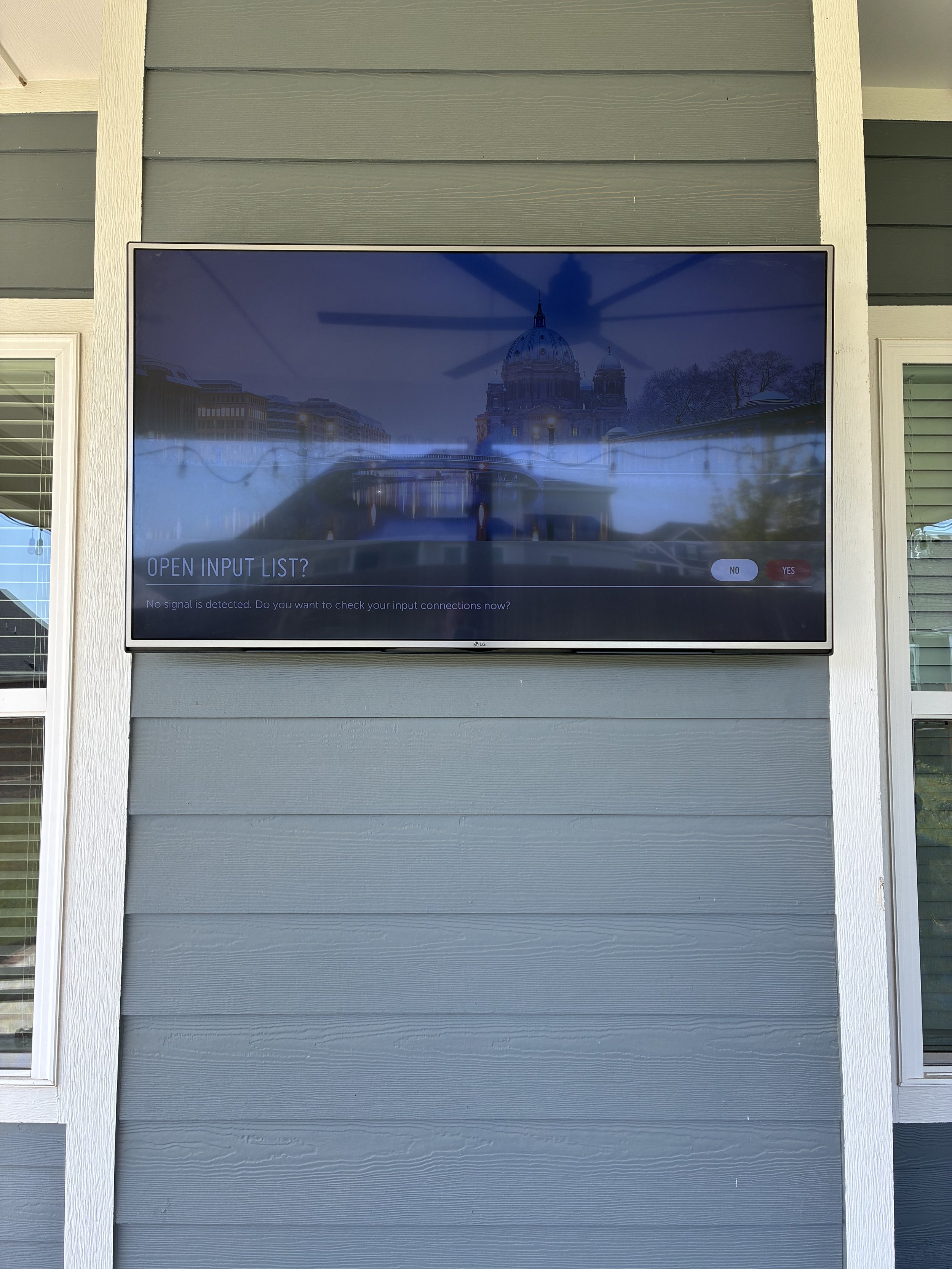 A flat screen TV mounted on a gray wooden wall showing a digital screen with the text 'OPEN INPUT LIST?' and options 'NO' and 'YES'. The display also shows a blurred reflection of a building with a dome and trees.
